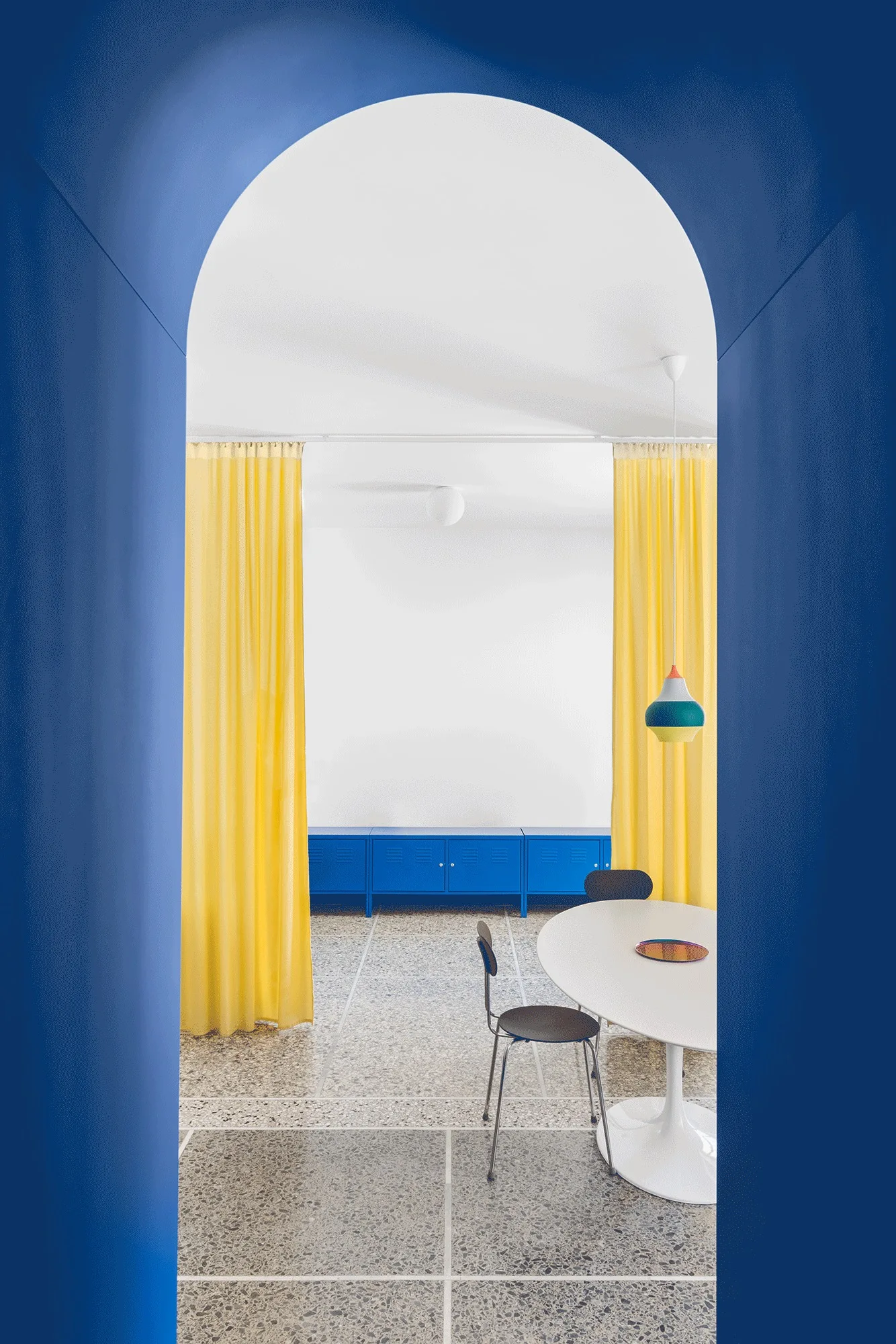 A view through a deep blue archway shows a dining area with a white pedestal table, bright yellow curtains, and a blue locker cabinet on terrazzo floors.