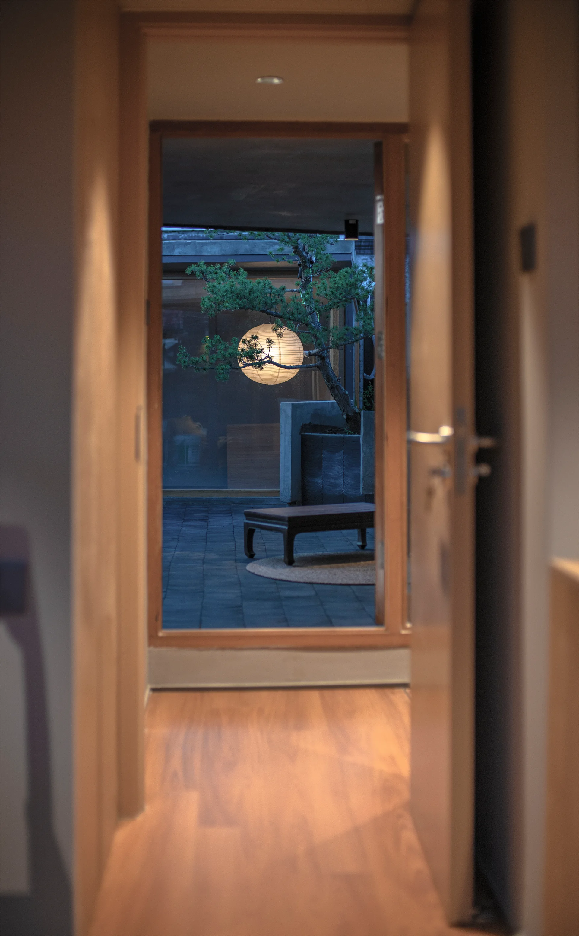 A view through an open wooden doorway looking onto a paved courtyard with a low wooden bench, a potted pine tree, and a glowing round paper lantern.
