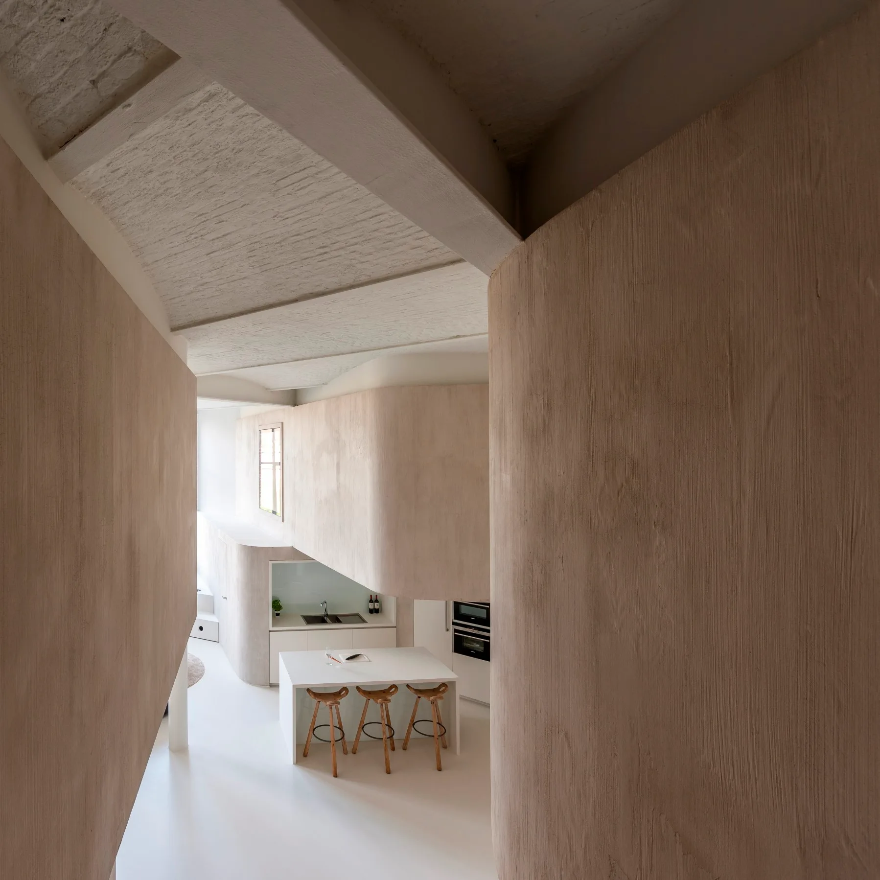 An elevated view of a minimalist kitchen featuring curved beige plaster walls, a white island with three wooden bar stools, and an exposed beam ceiling.