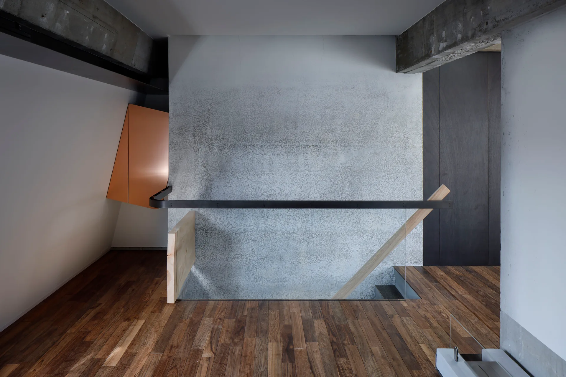 A minimalist modern stairwell features a textured concrete wall, dark wood floors, a black metal handrail, and concrete ceiling beams.