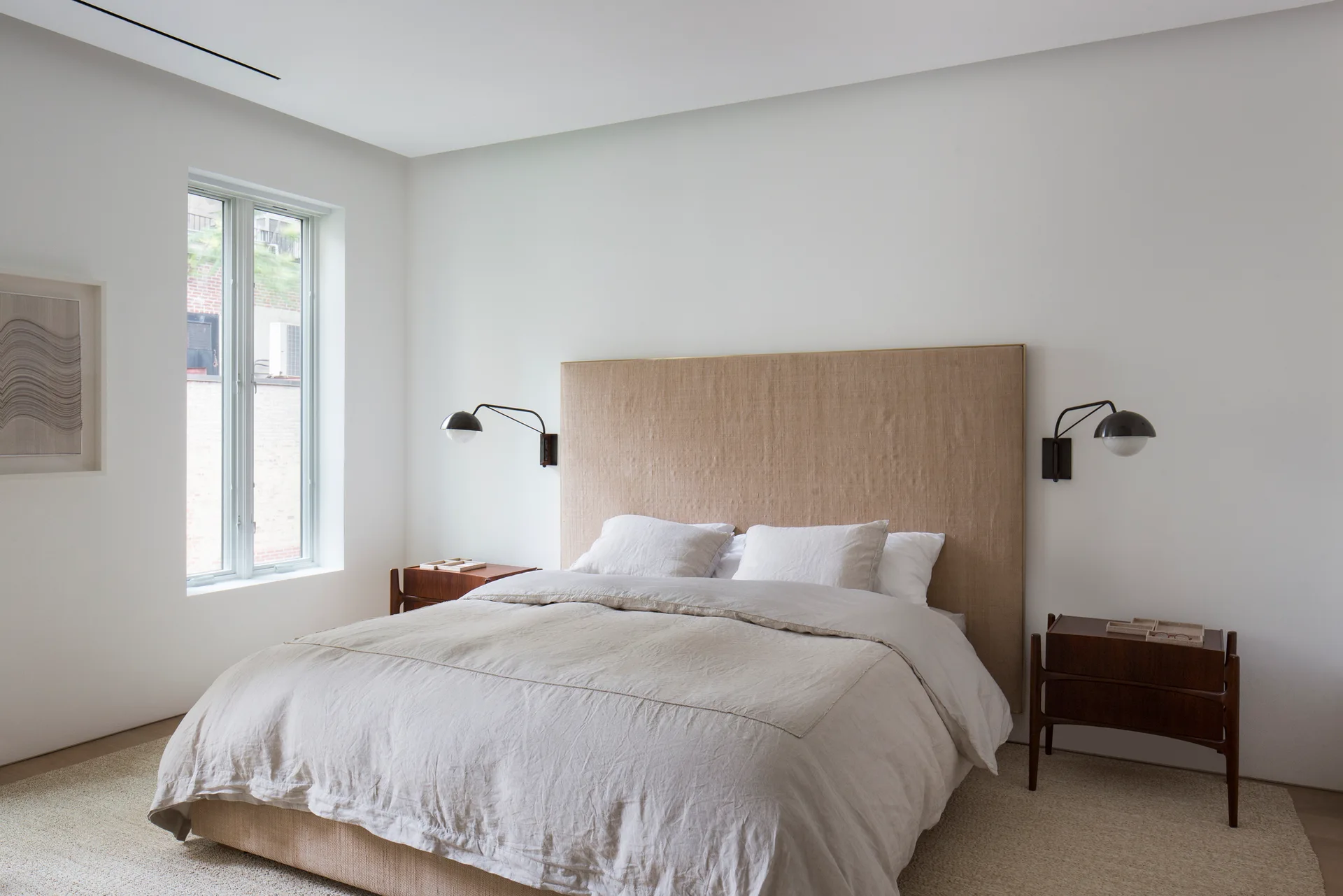 A minimalist bedroom featuring a tall tan linen headboard, dark wood nightstands, black wall sconces, and neutral bedding on a textured rug.