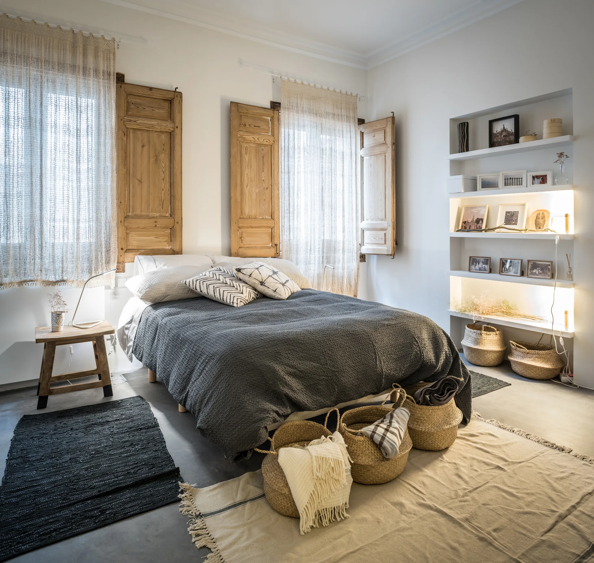 A rustic bedroom features a bed with a dark grey blanket, warm wood window shutters, and white recessed wall shelves with integrated lighting.