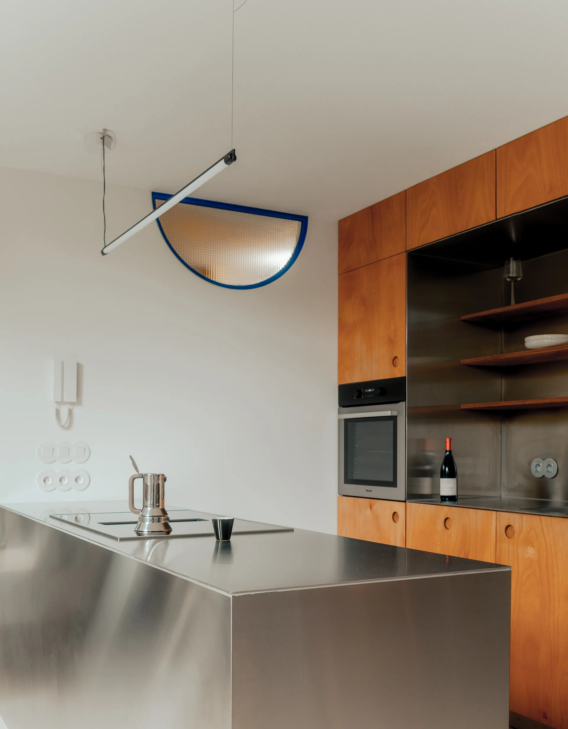 A modern kitchen featuring a stainless steel island, warm wood cabinetry, a linear pendant light, and a semi-circular wall cutout.