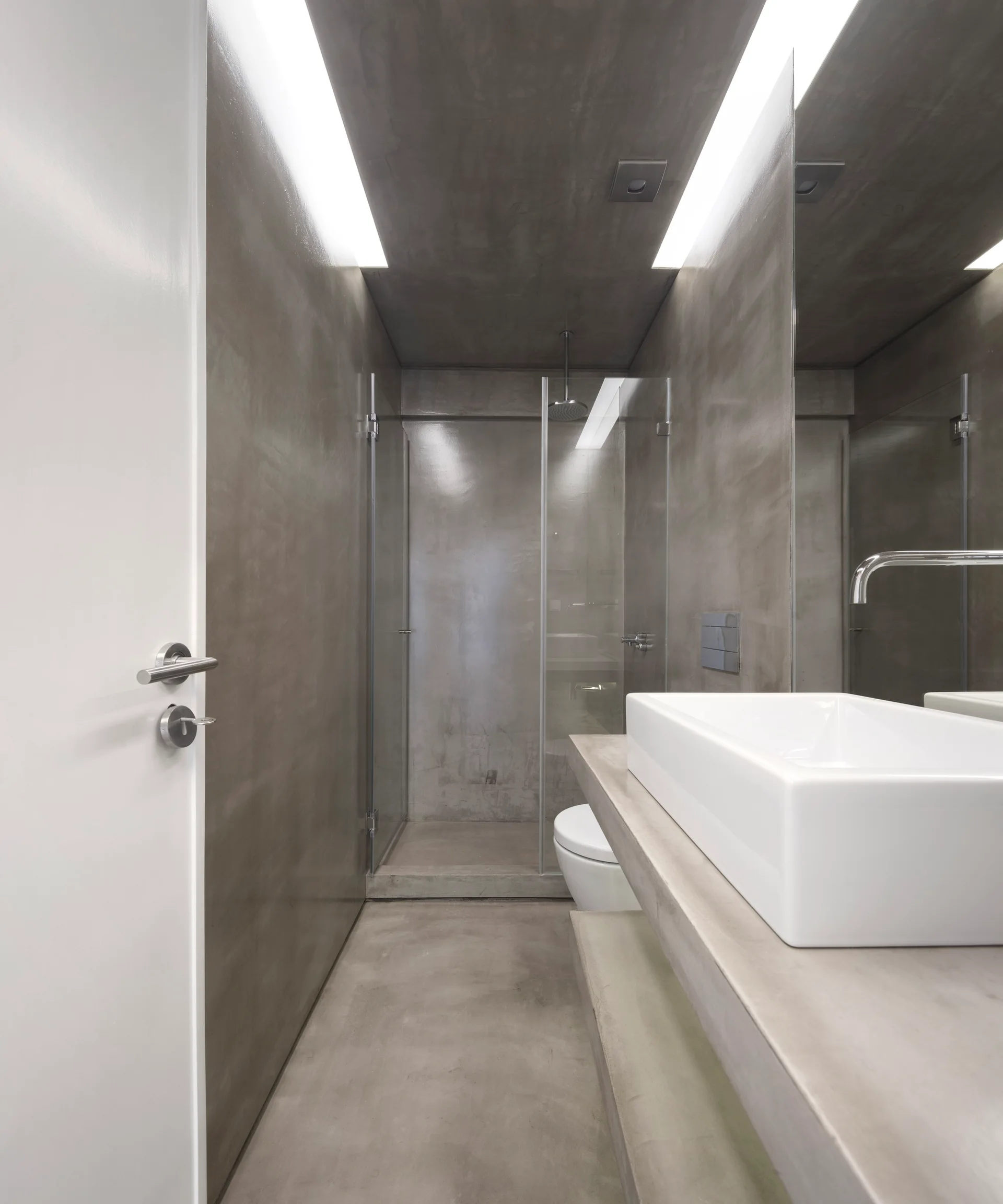 A minimalist grey concrete bathroom featuring a white rectangular vessel sink, wall-mounted mirror, and linear cove lighting along the ceiling.