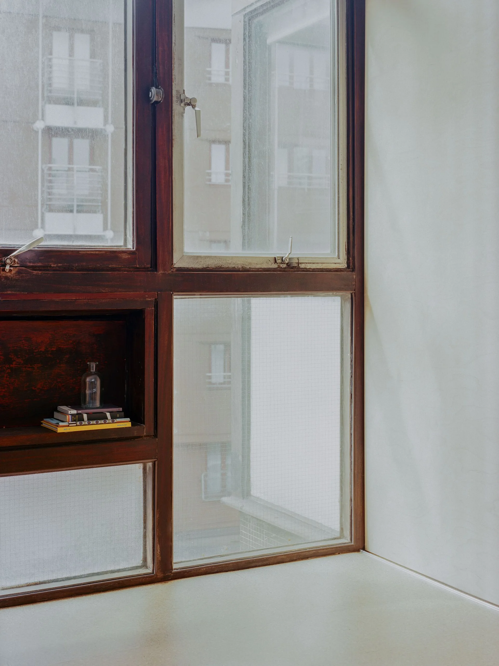 A dark brown wooden window frame features a built-in display shelf holding a stack of books and a small glass bottle next to a plain off-white wall.