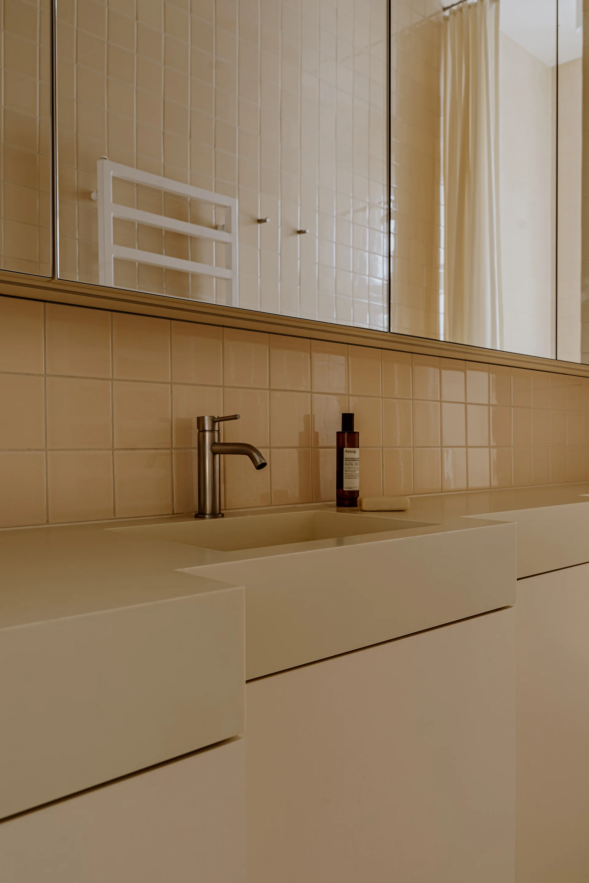 A minimalist beige bathroom vanity with an integrated sink, a brushed metal faucet, and a square ceramic tile backsplash beneath a mirrored cabinet.