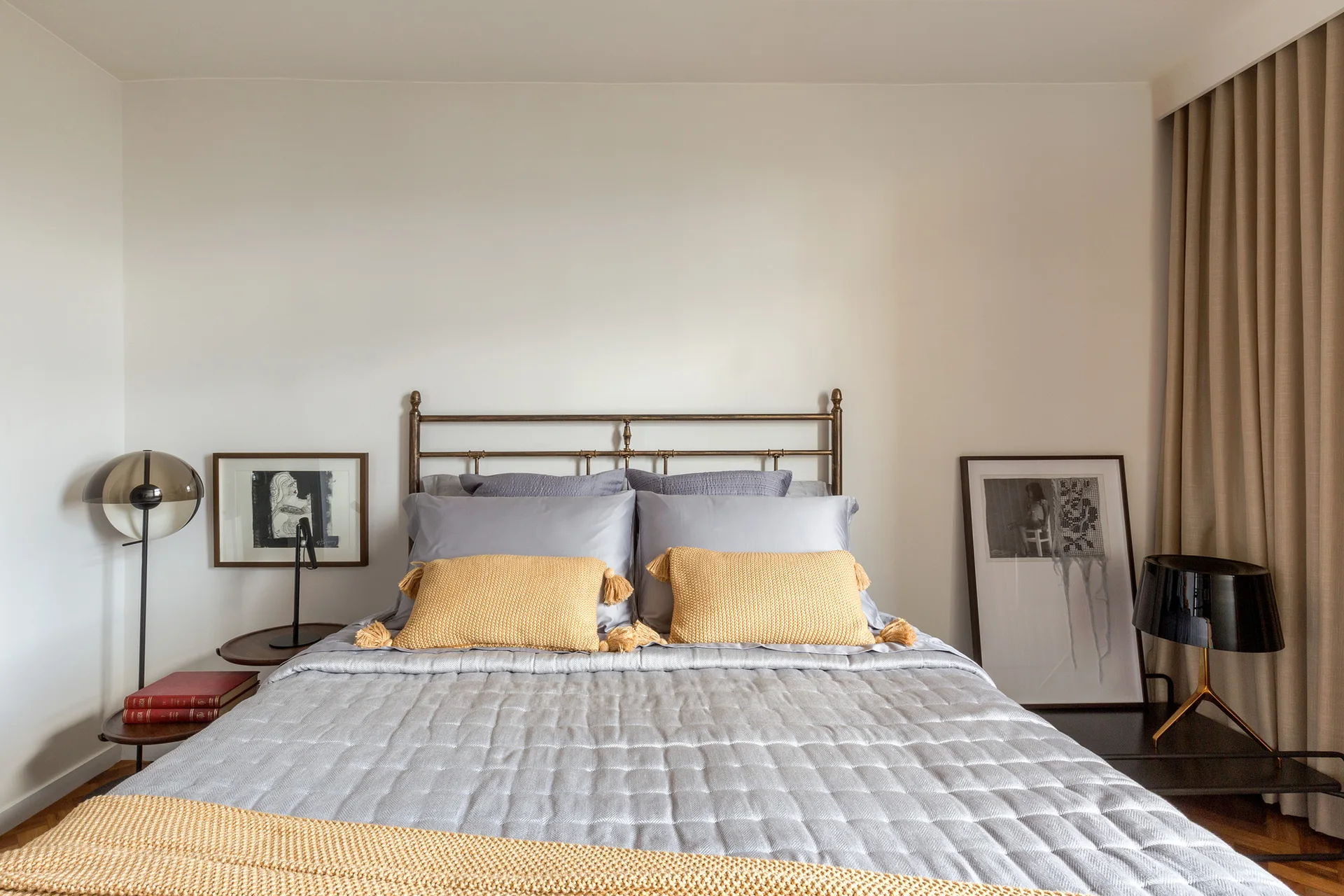 A metal frame bed with quilted grey bedding and mustard yellow tasseled pillows sits between two modern bedside tables with lamps.