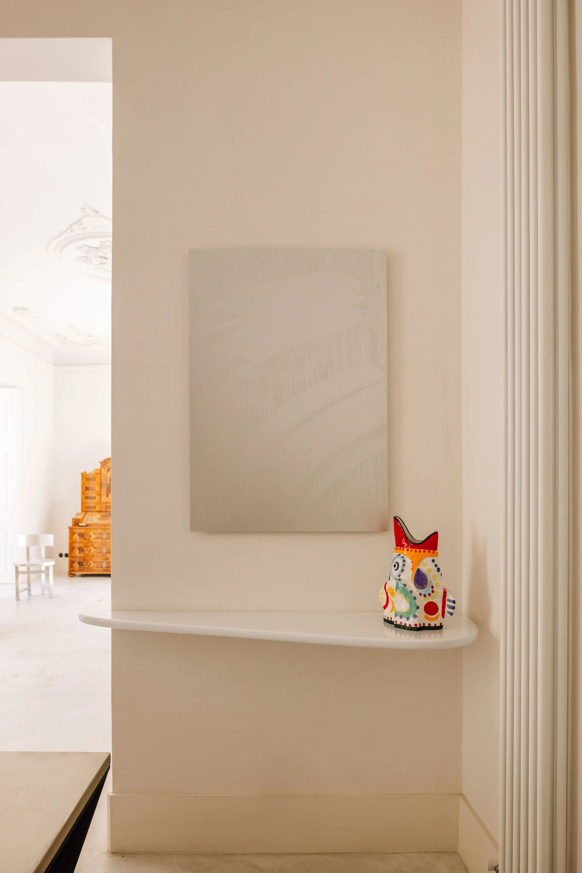A curved white floating shelf mounted on an off-white wall holds a colorful patterned ceramic bird sculpture below a textured minimalist canvas.