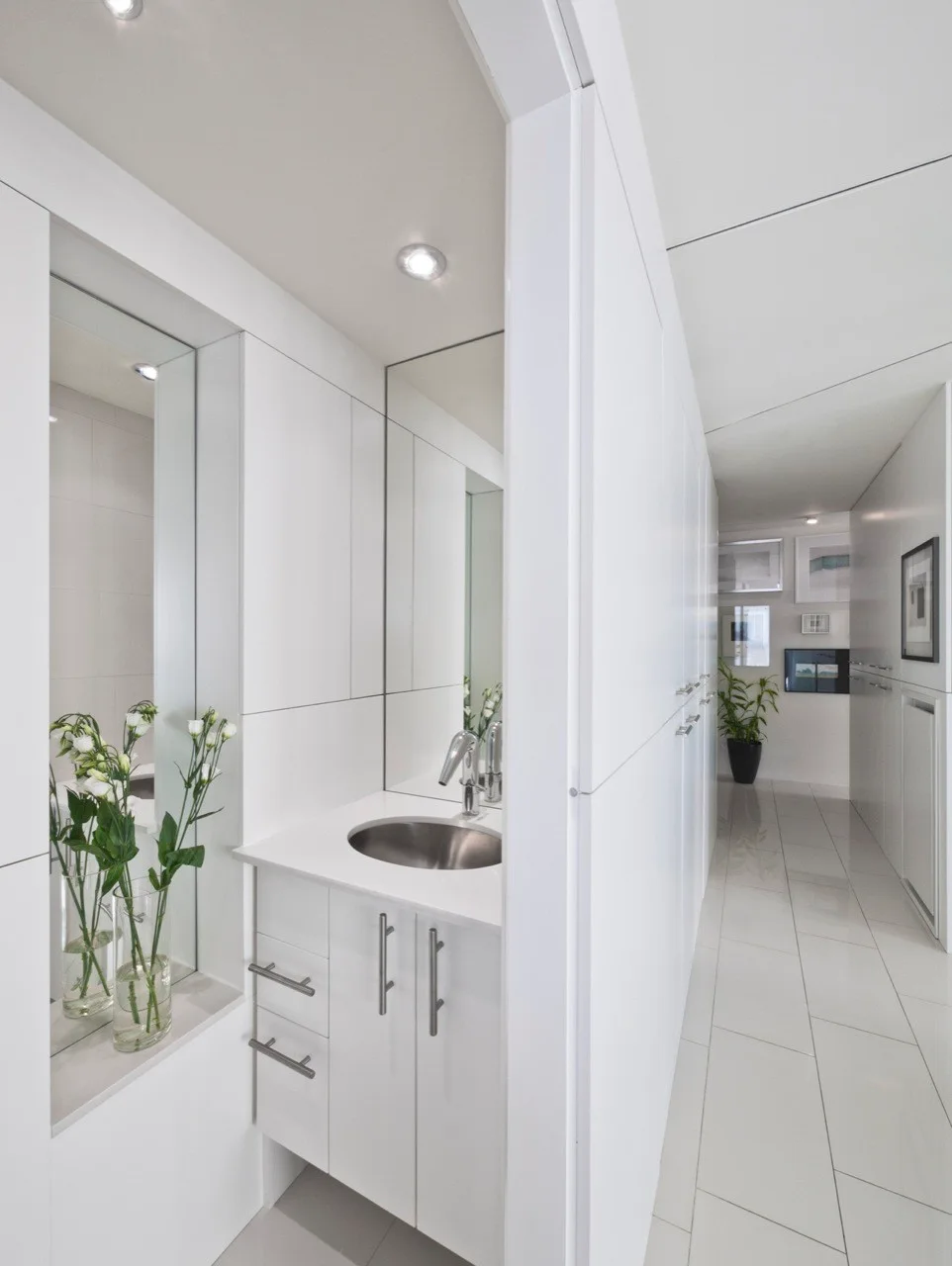 A minimalist white vanity featuring an integrated stainless steel sink and a recessed wall mirror beside a bright tiled hallway.