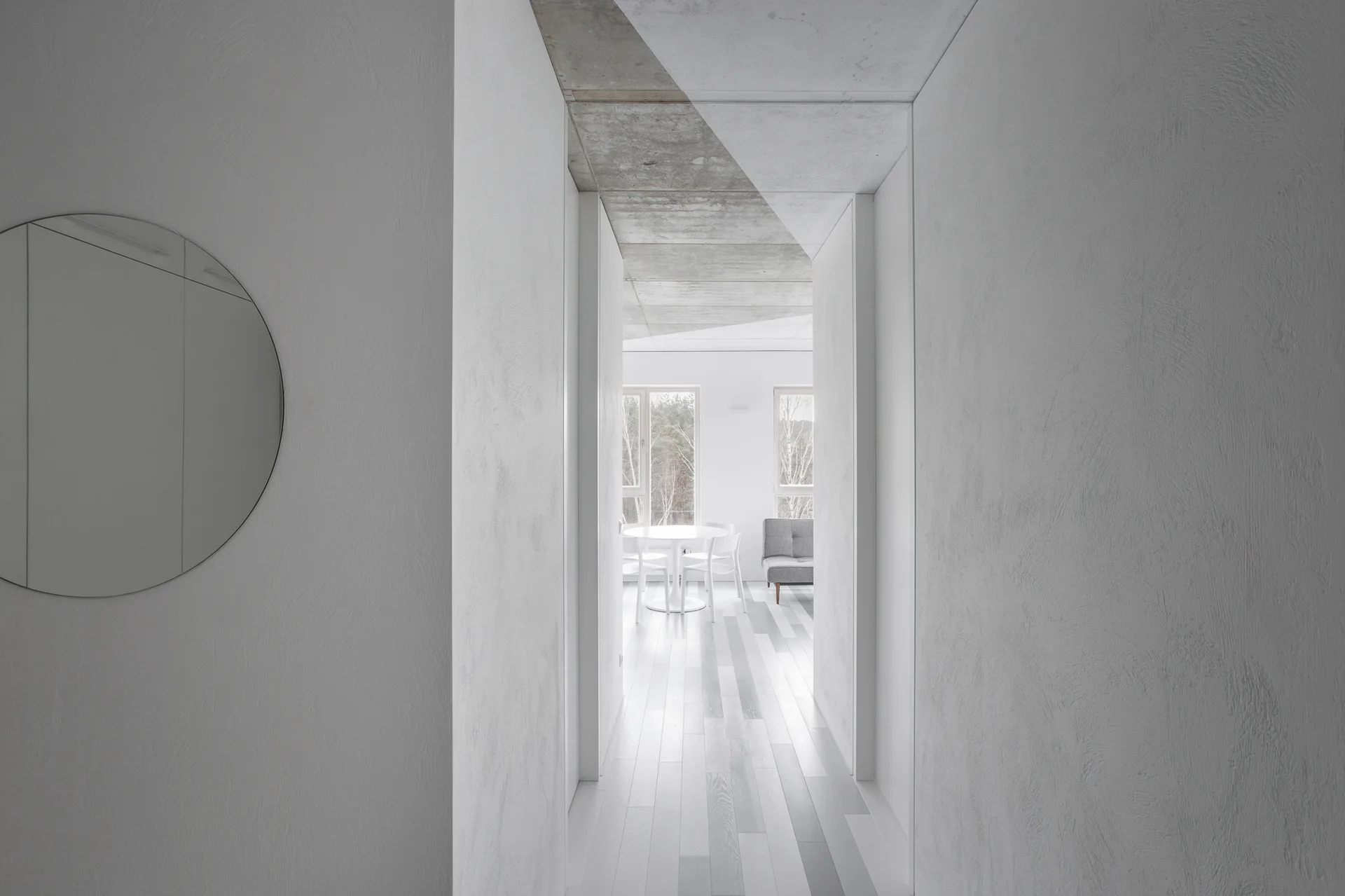 A minimalist hallway with textured white plaster walls, a round mirror, and an exposed concrete ceiling looking into a bright dining area.