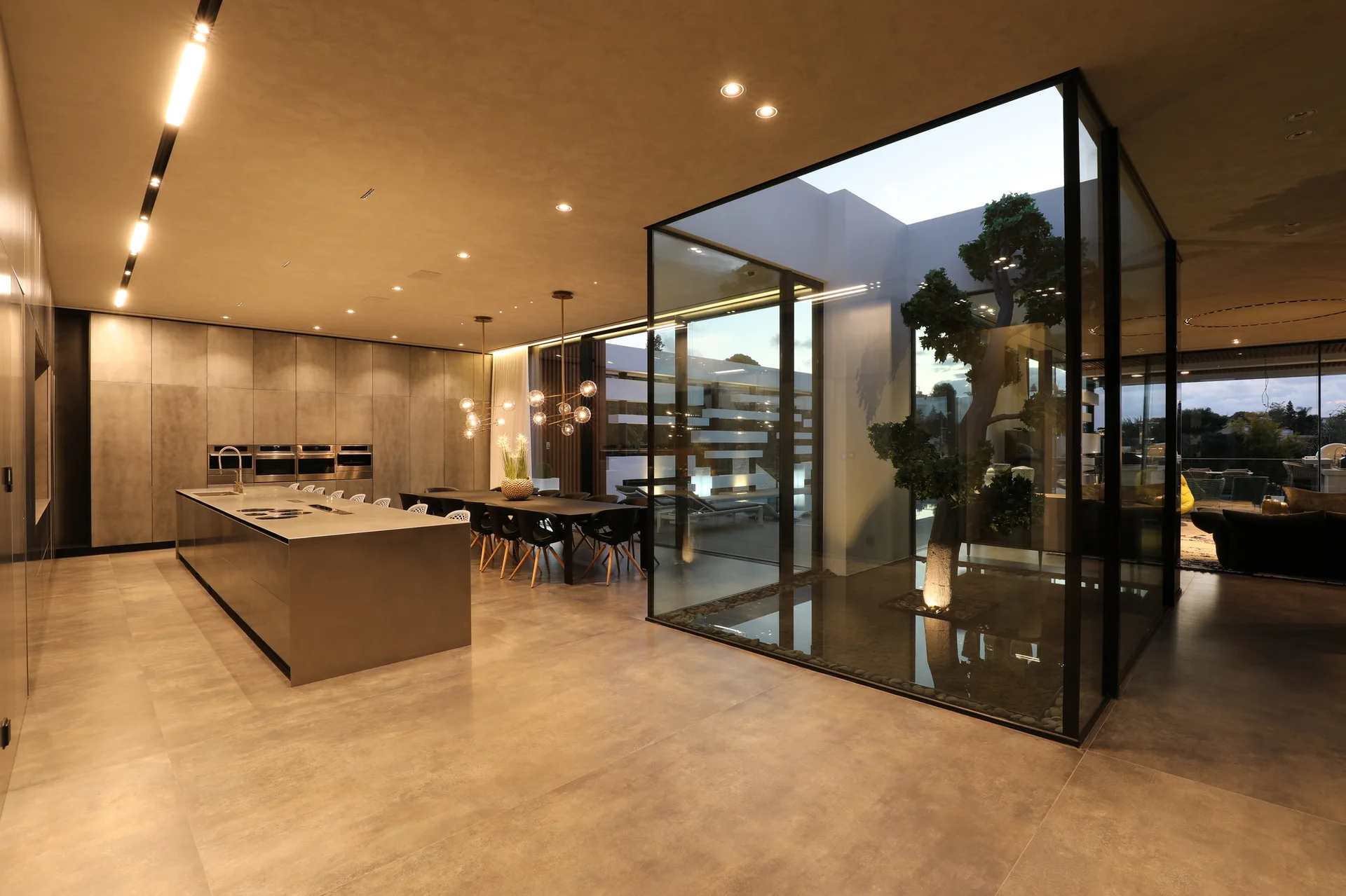 A minimalist kitchen and dining space featuring a large metallic island, flat-front cabinetry, and a central glass atrium with an indoor tree.