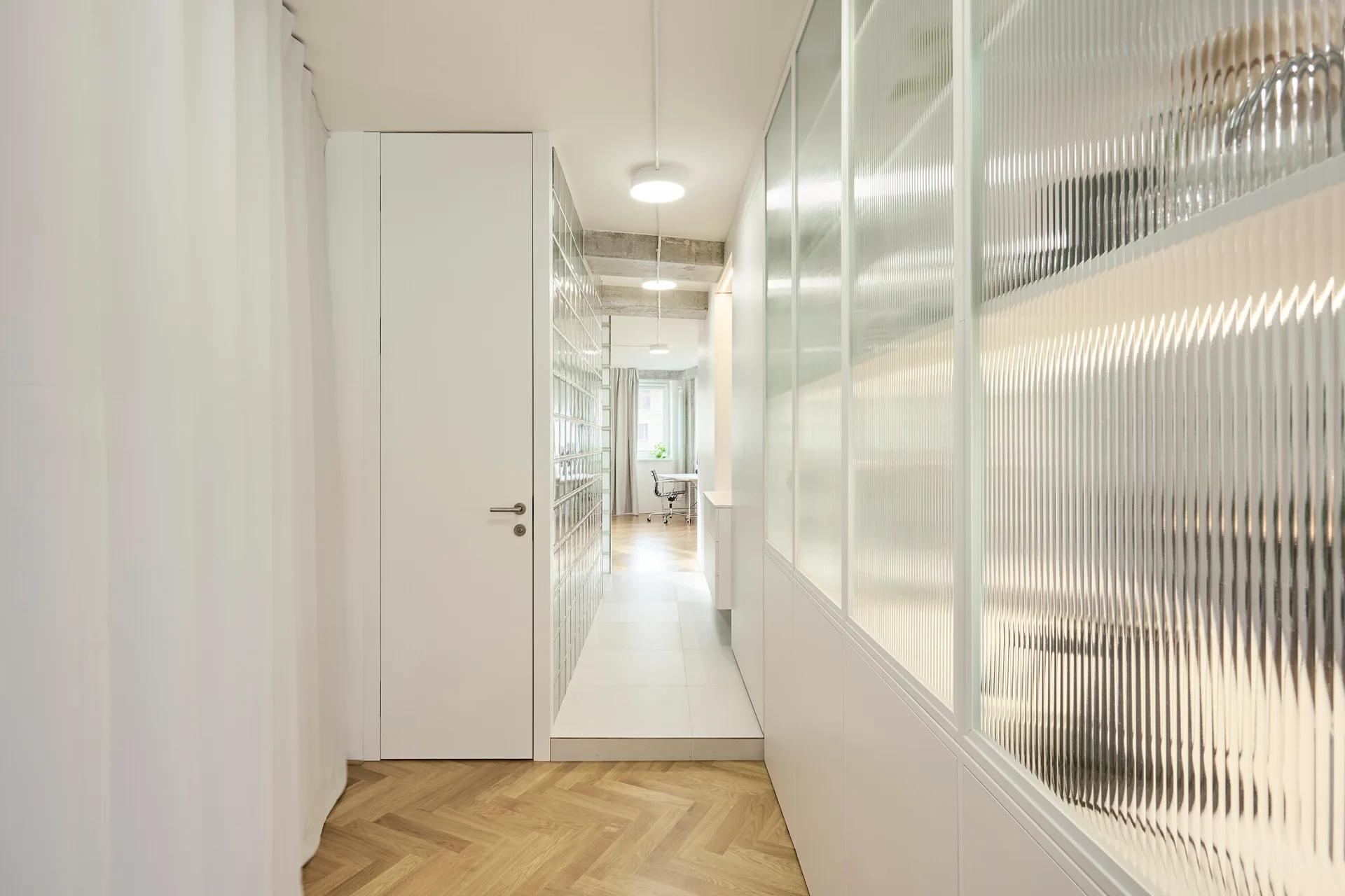 A minimalist white hallway featuring herringbone wood flooring, a fluted glass partition wall, and integrated storage cabinets.
