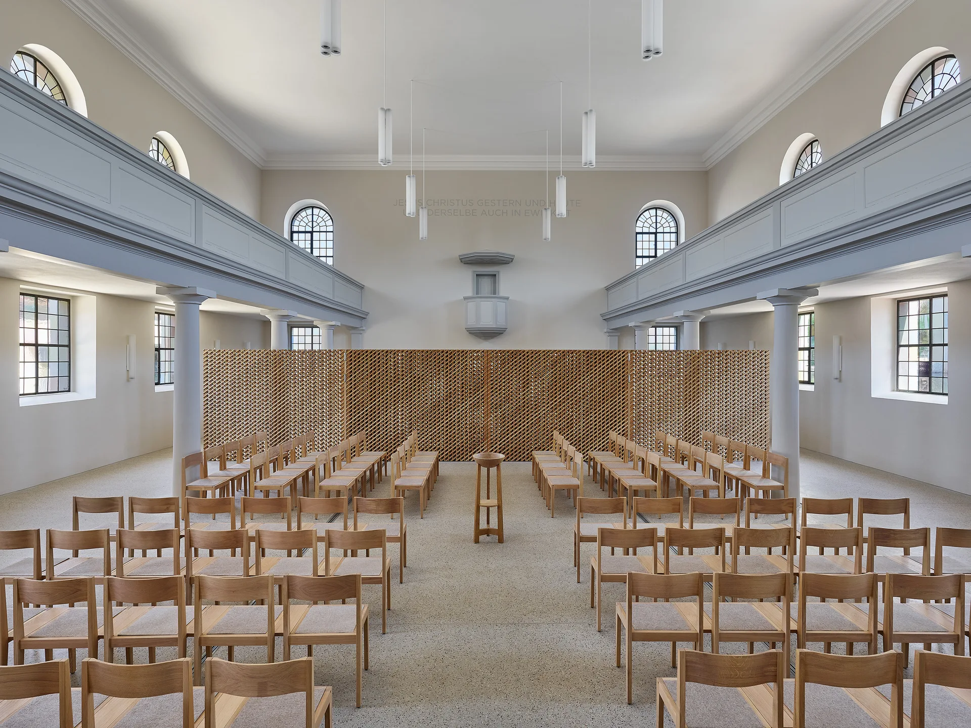Rows of light wood chairs face a large wooden lattice screen inside a symmetrical hall with columns, upper balconies, and arched windows.