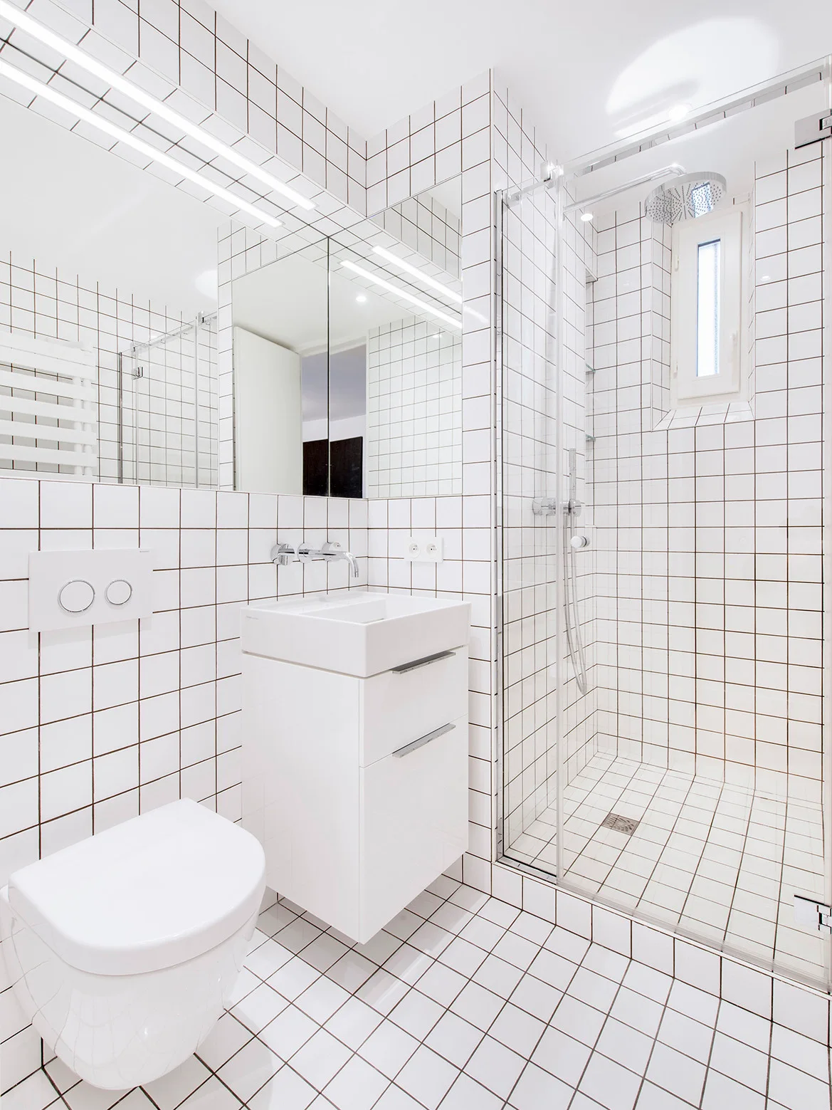 A minimalist white bathroom features square grid tiling on the walls and floor, a wall-mounted vanity, and a glass-enclosed shower.