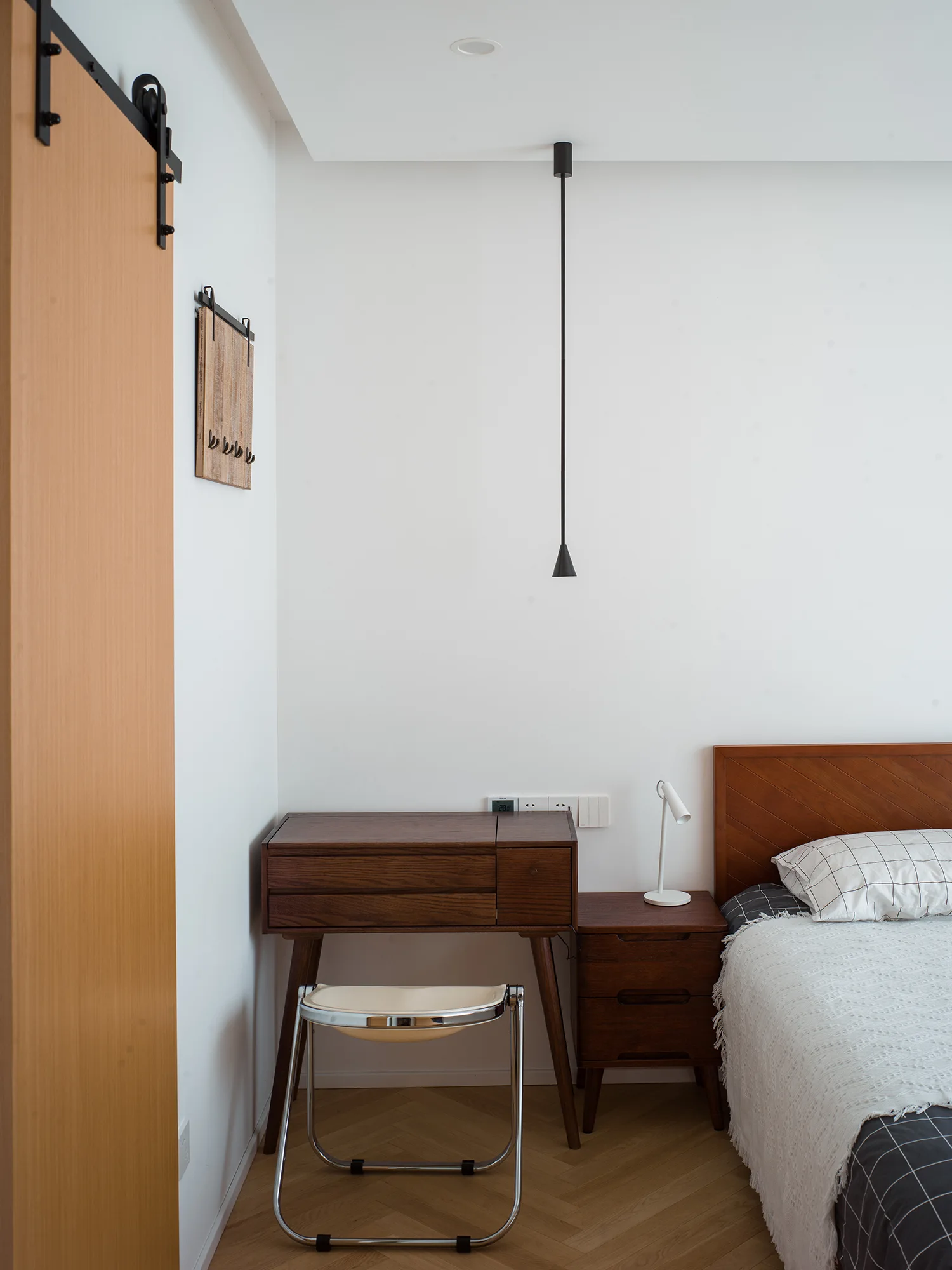 A minimalist bedroom features a dark wood bed and writing desk, a white task lamp, a black pendant light, and a light wood sliding barn door.