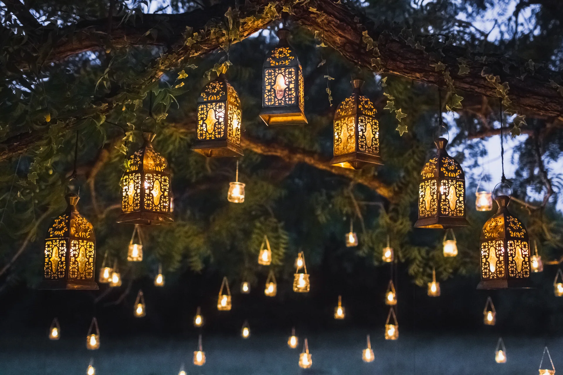 Several ornate metal lanterns and small glass lights hang glowing from tree branches at night.