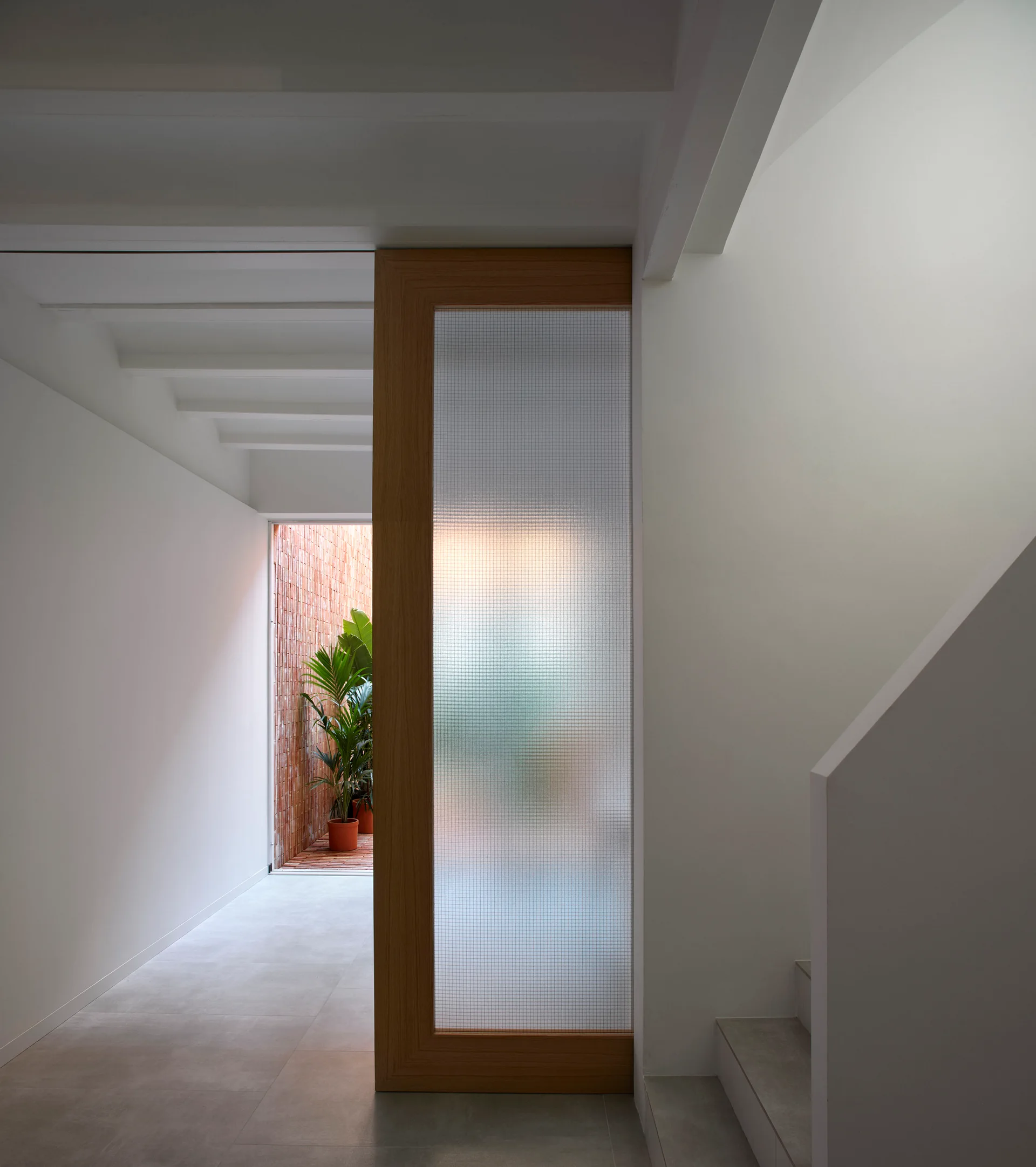 A minimalist white hallway features a tall wood-framed sliding door with textured glass next to a staircase and a view of a small brick courtyard.