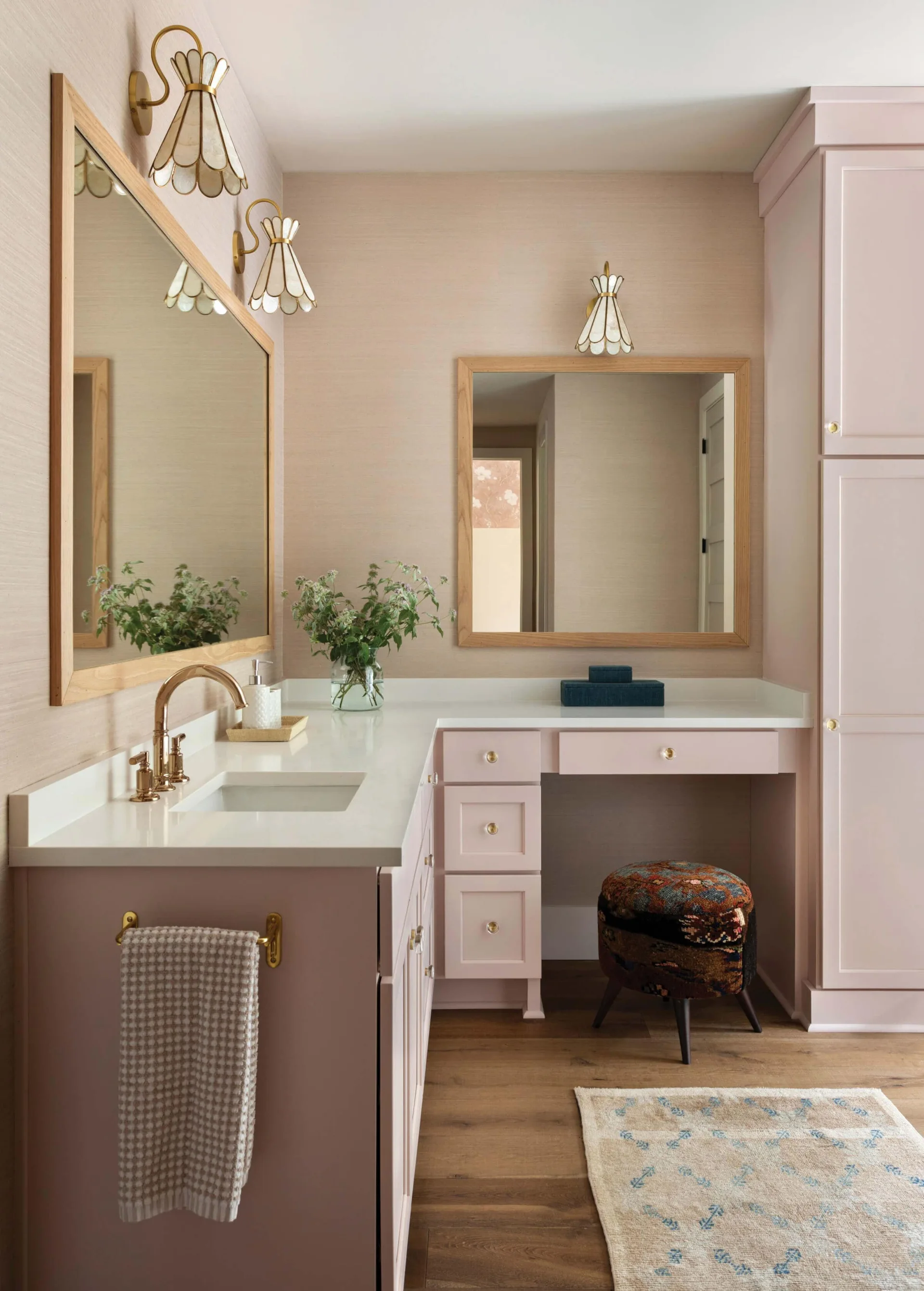 A bathroom features a custom blush pink vanity with a white countertop, gold faucet, wood-framed mirrors, and a patterned stool.