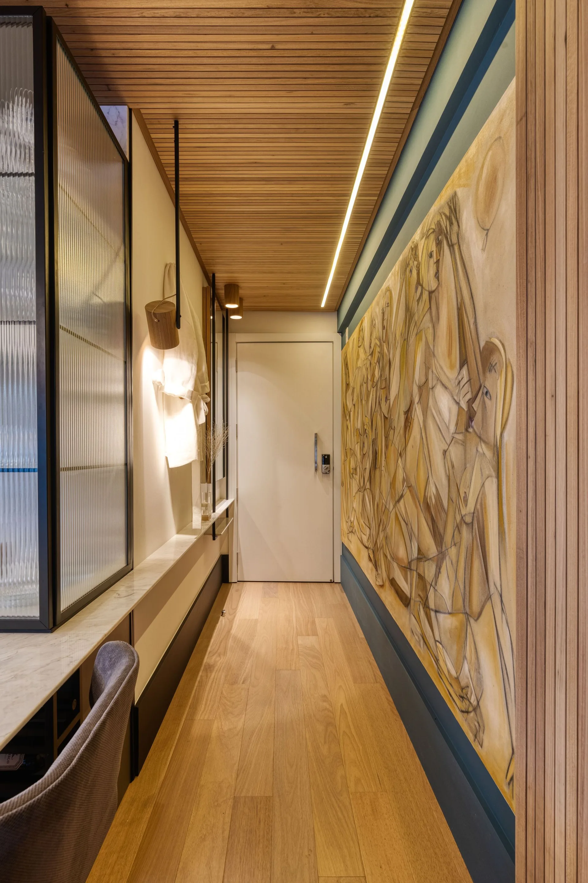 A narrow residential hallway features a light wood floor, a wood-slatted ceiling with linear LED lighting, and a large painting on the right wall.