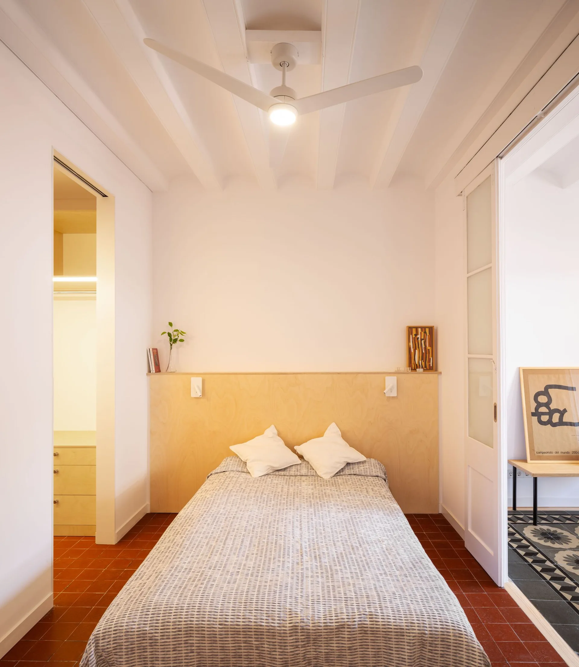 A minimalist bedroom features a bed with a light wood plywood headboard, red tile flooring, white beamed ceilings, and an adjacent lighted closet.