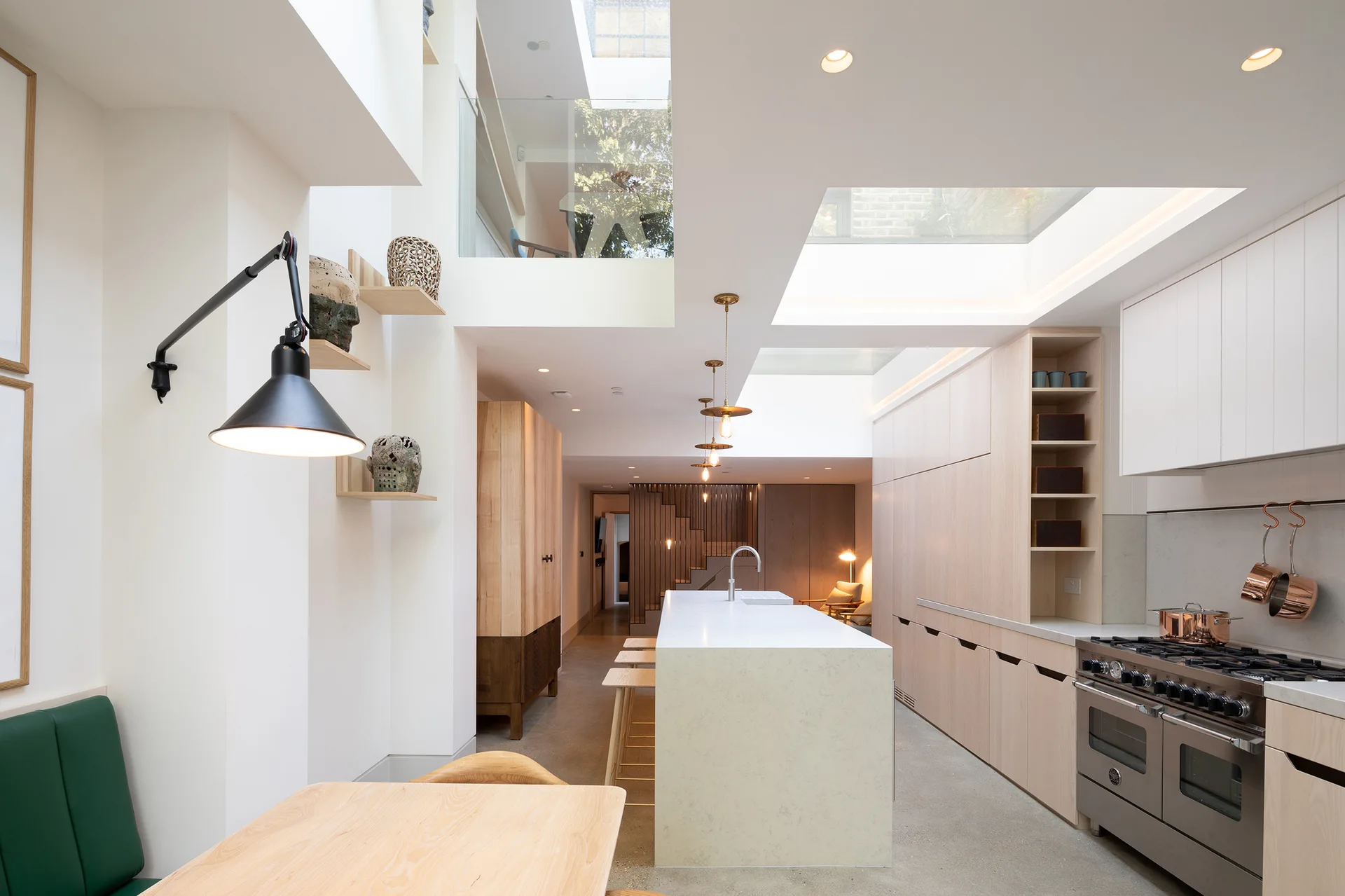 A modern kitchen features light wood cabinets, a long white stone-topped island, concrete floors, and large overhead skylights.