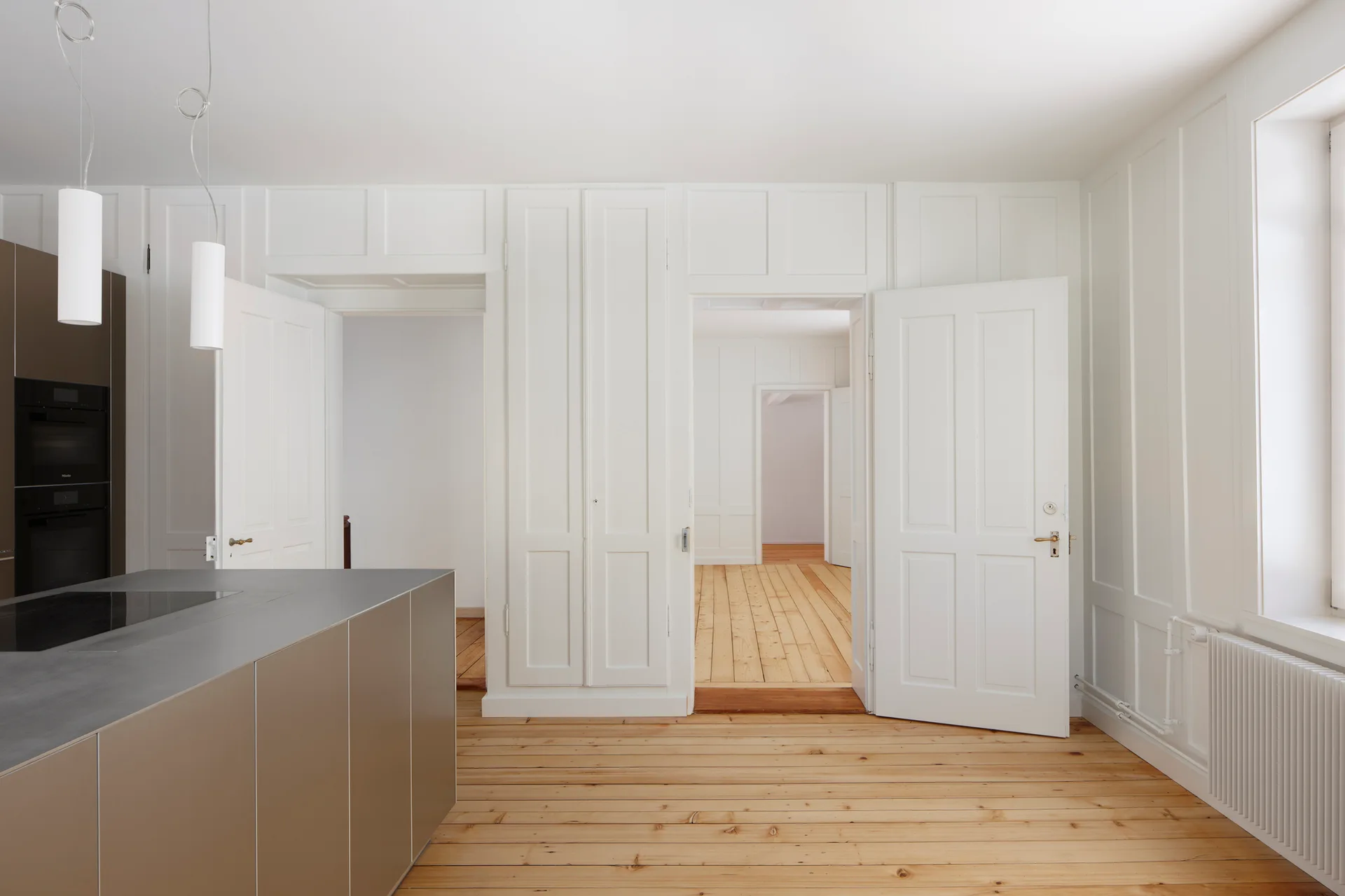 A modern metallic kitchen island with a dark cooktop sits on light wood floors in a room with white paneled walls and open doors.