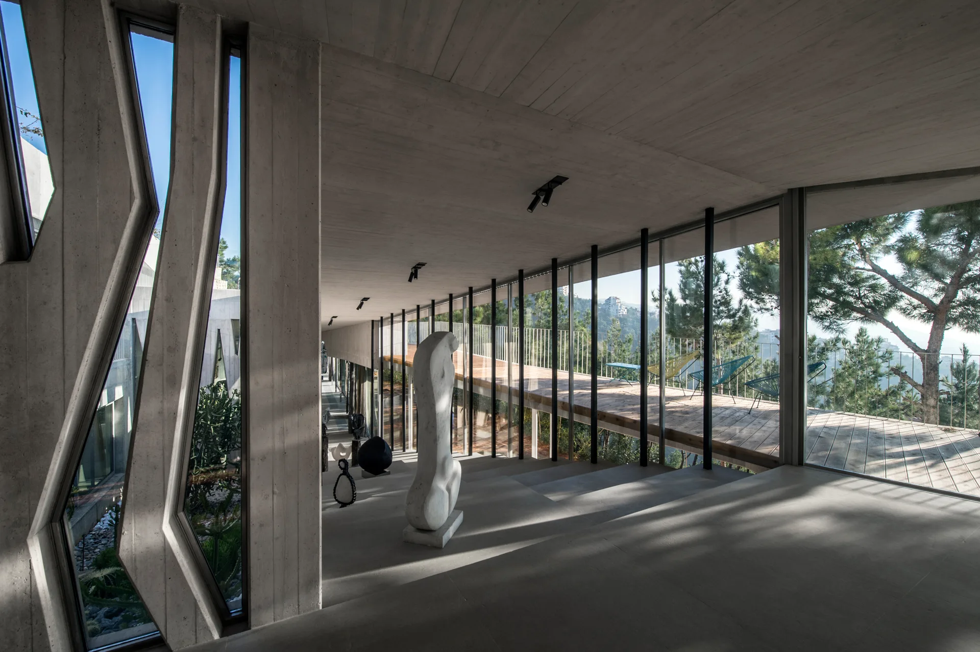 A long minimalist concrete corridor features floor-to-ceiling glass walls, a smooth grey floor, and abstract sculptures displayed along the path.