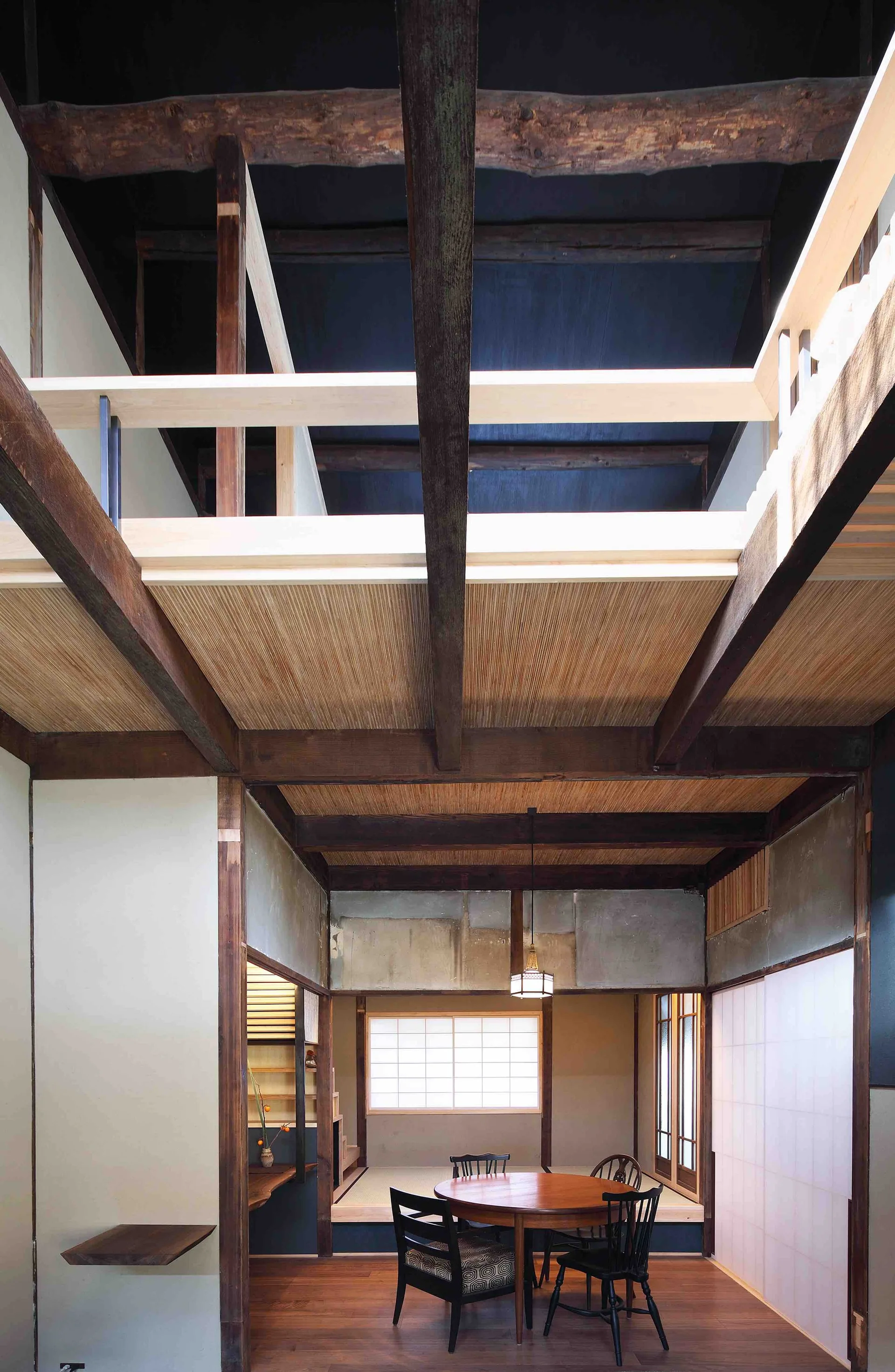 A Japanese style dining room with a round wooden table, black chairs, a raised tatami mat platform, and high ceilings with exposed wood beams.