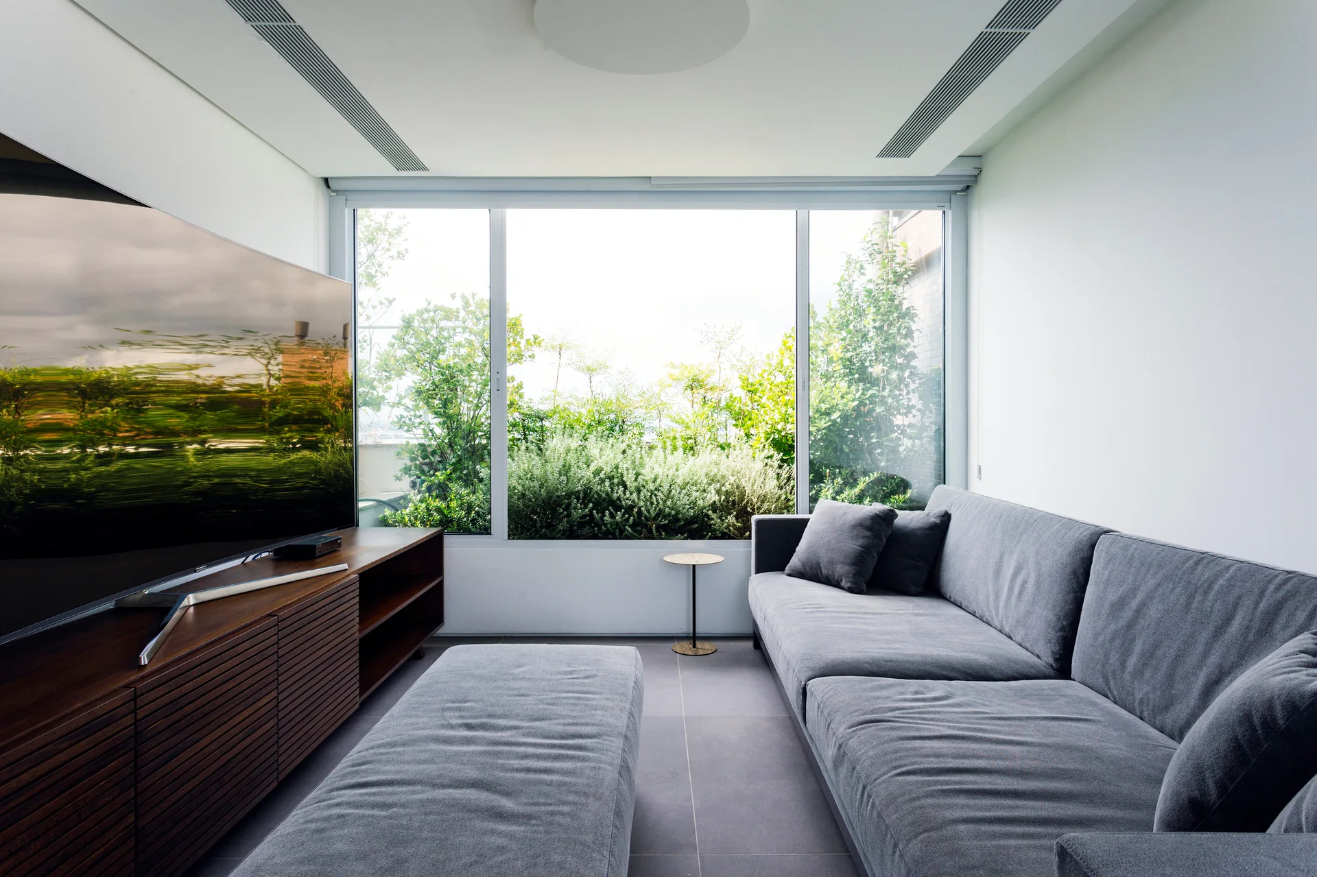 A minimalist living room with a grey sectional sofa, slatted wood media console, and a large picture window showcasing lush green outdoor plants.
