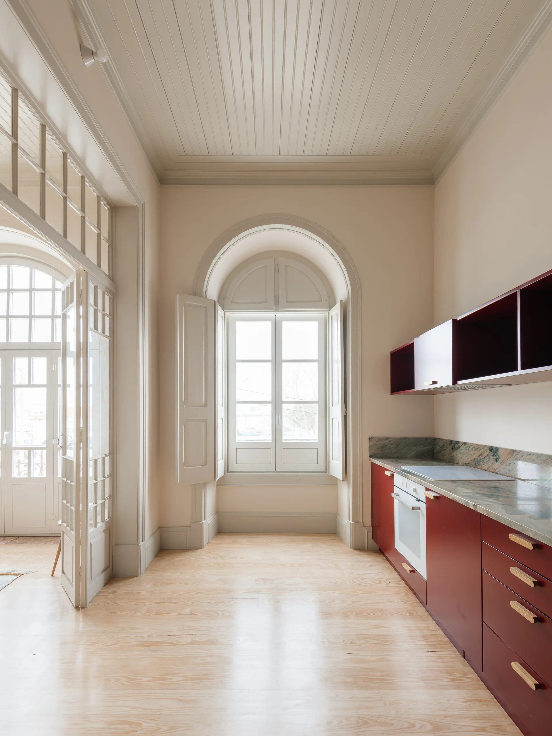 A minimalist kitchen features deep red cabinetry, a stone countertop, light wood flooring, beige walls, and a tall arched window.