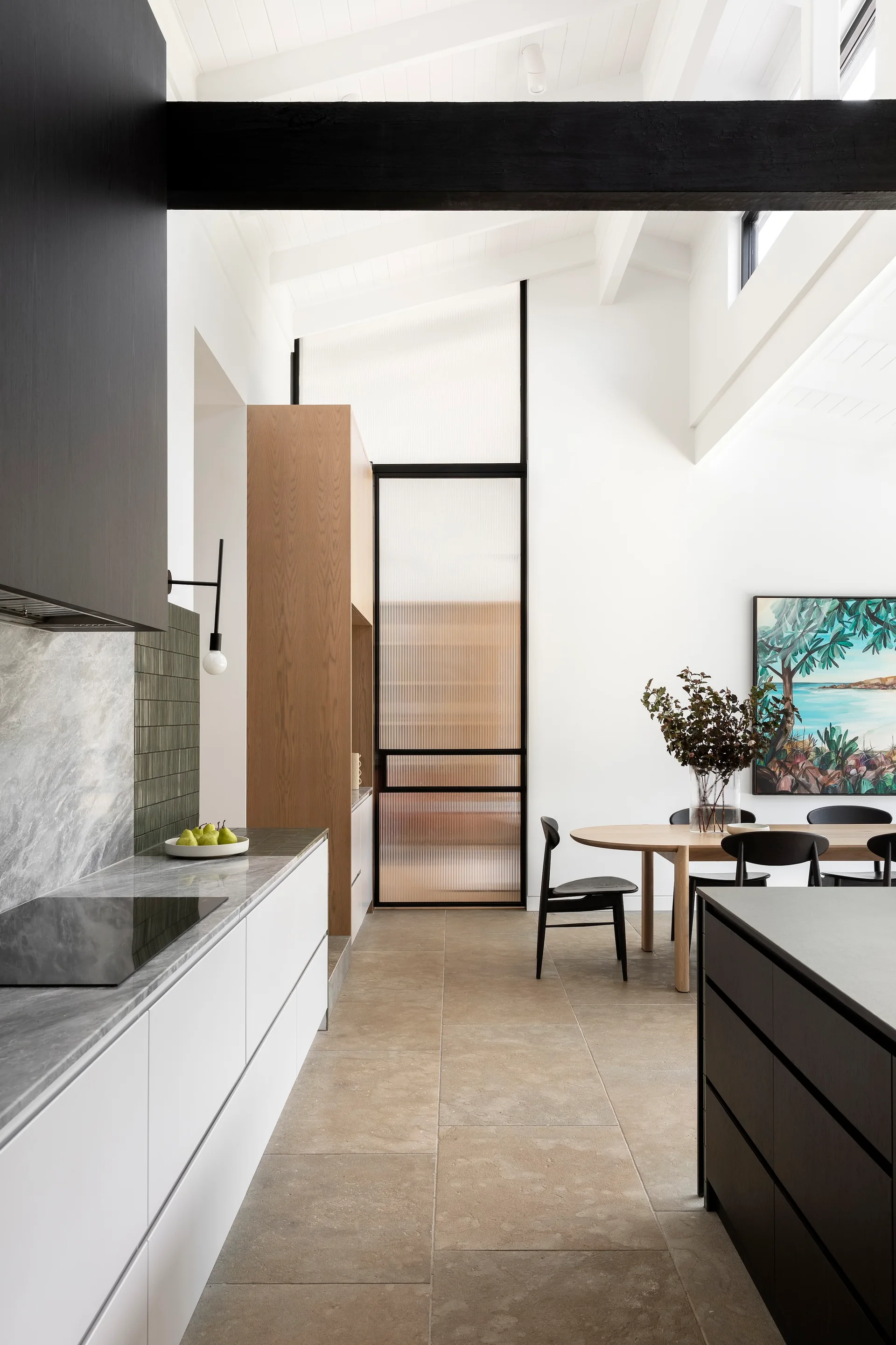A modern kitchen and dining space with white cabinets, dark wood beams, and a fluted glass door under a high vaulted ceiling.