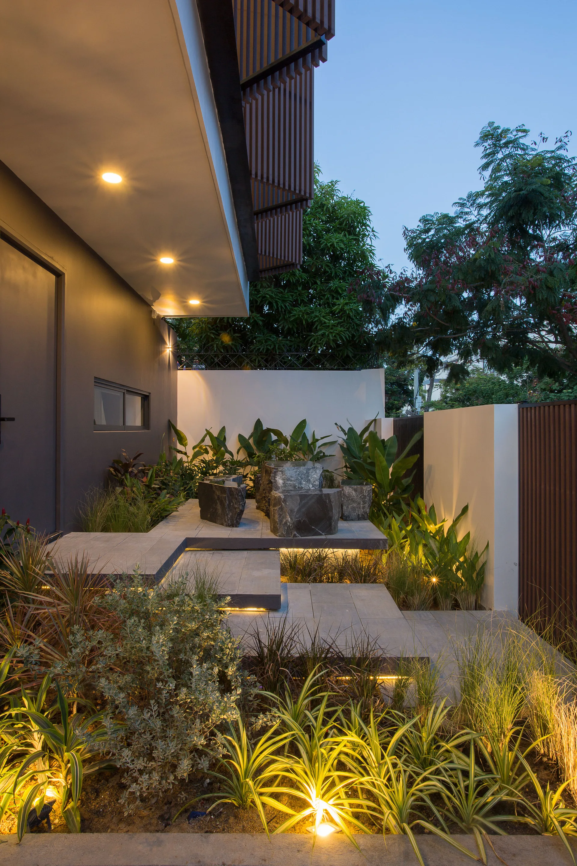 A modern tiered concrete patio featuring sculptural dark stone block seating surrounded by lush green landscaping and warm architectural step lighting.