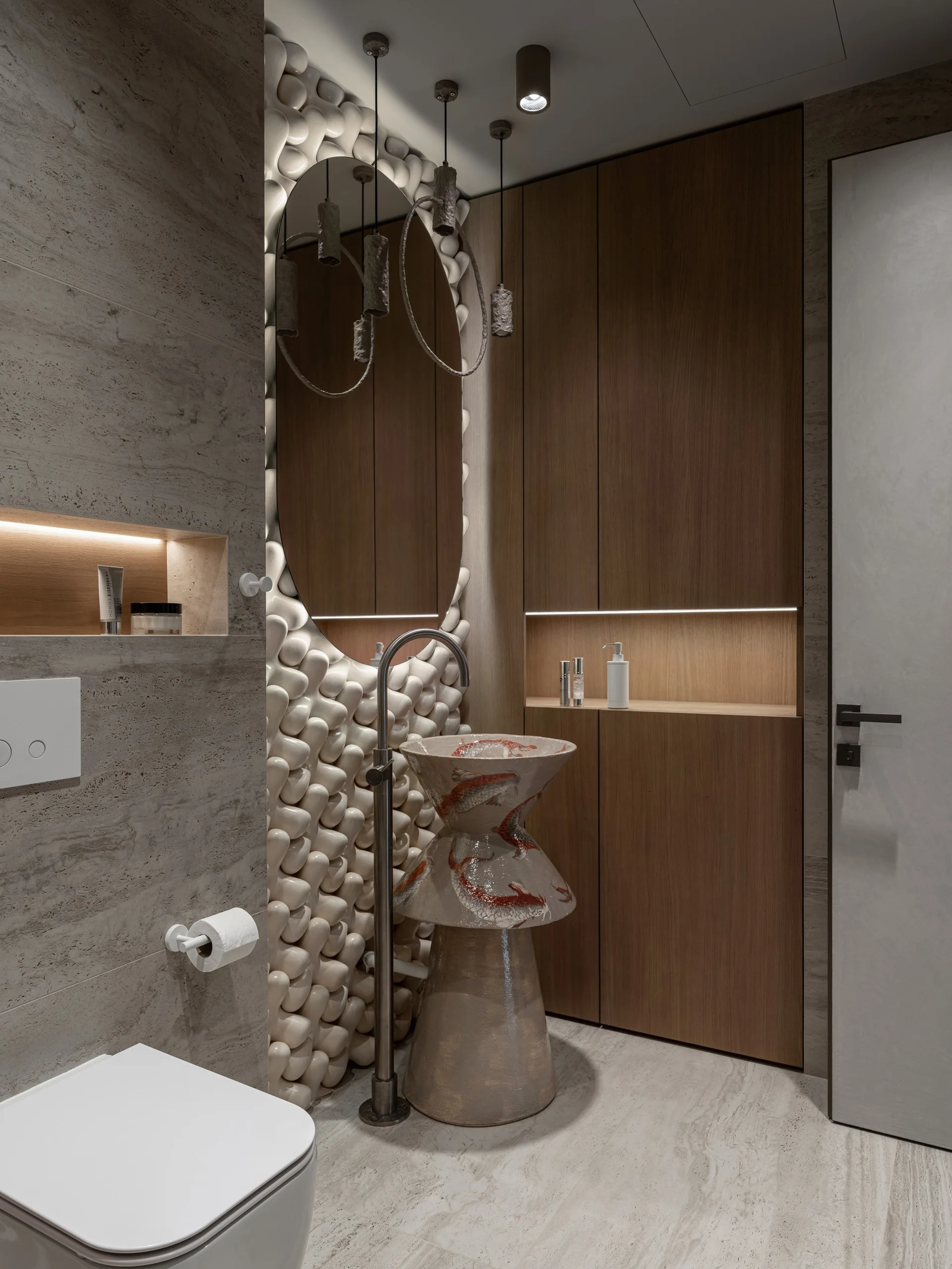 A modern powder room features a custom ceramic pedestal sink, an oval mirror on a highly textured white wall, and tall wood cabinetry with a lit niche.