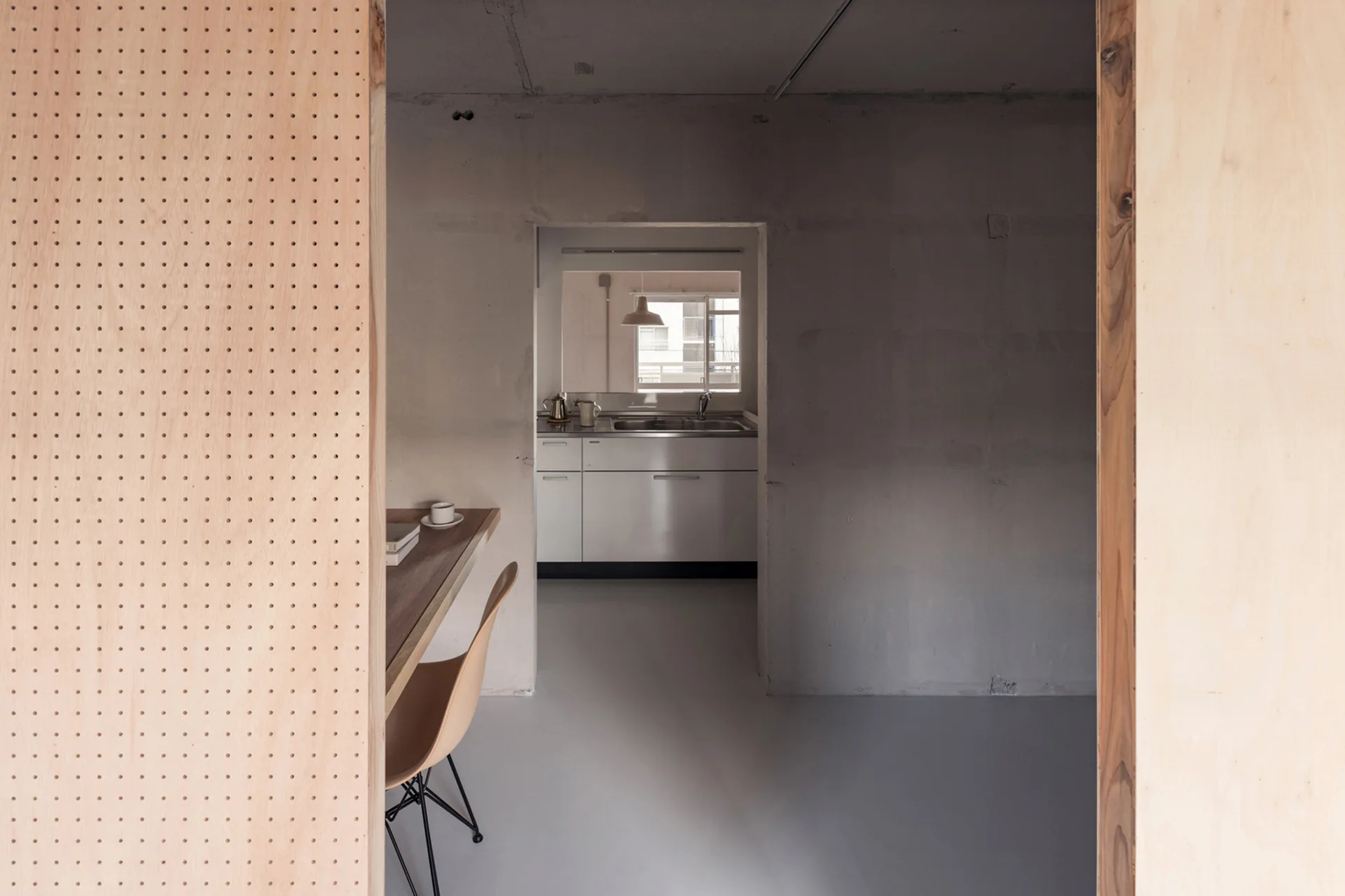 A light wood pegboard partition frames a view of a built-in study desk leading to a minimalist kitchen with grey walls and white cabinets.