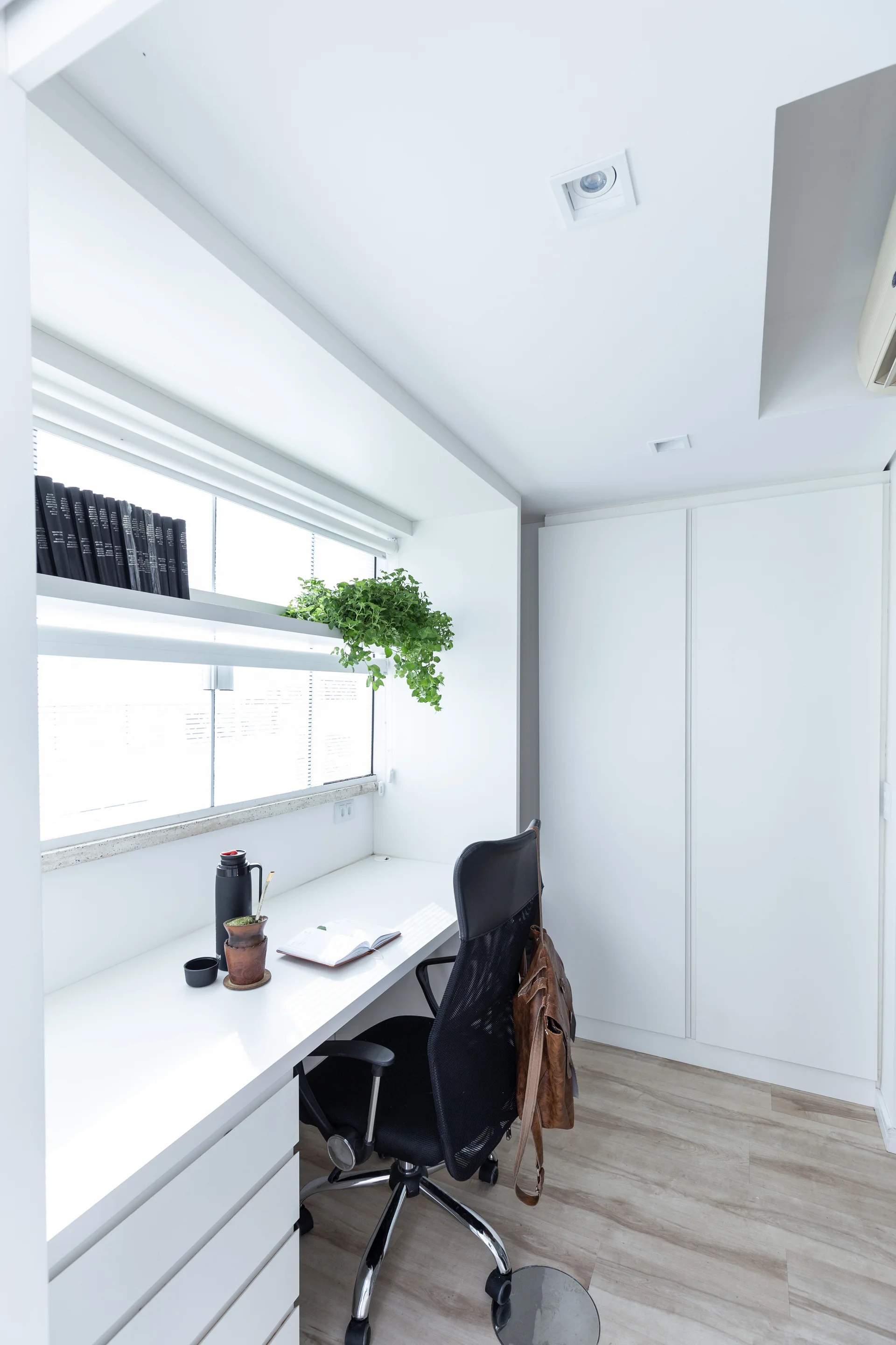 A minimalist home office featuring a white built-in desk under a window with a black office chair and built-in white cabinets.