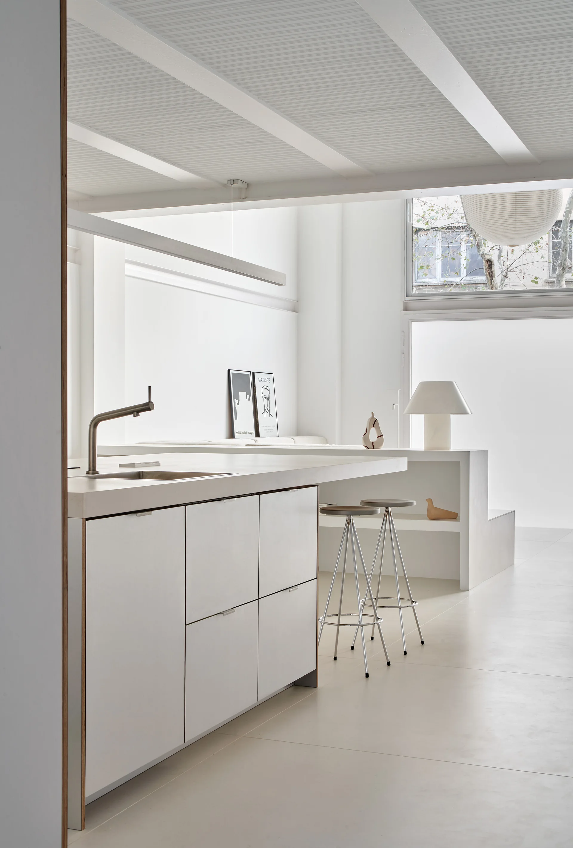 A minimalist white kitchen features a matte finish island with bar stools, an integrated low storage shelf, and exposed painted ceiling beams.