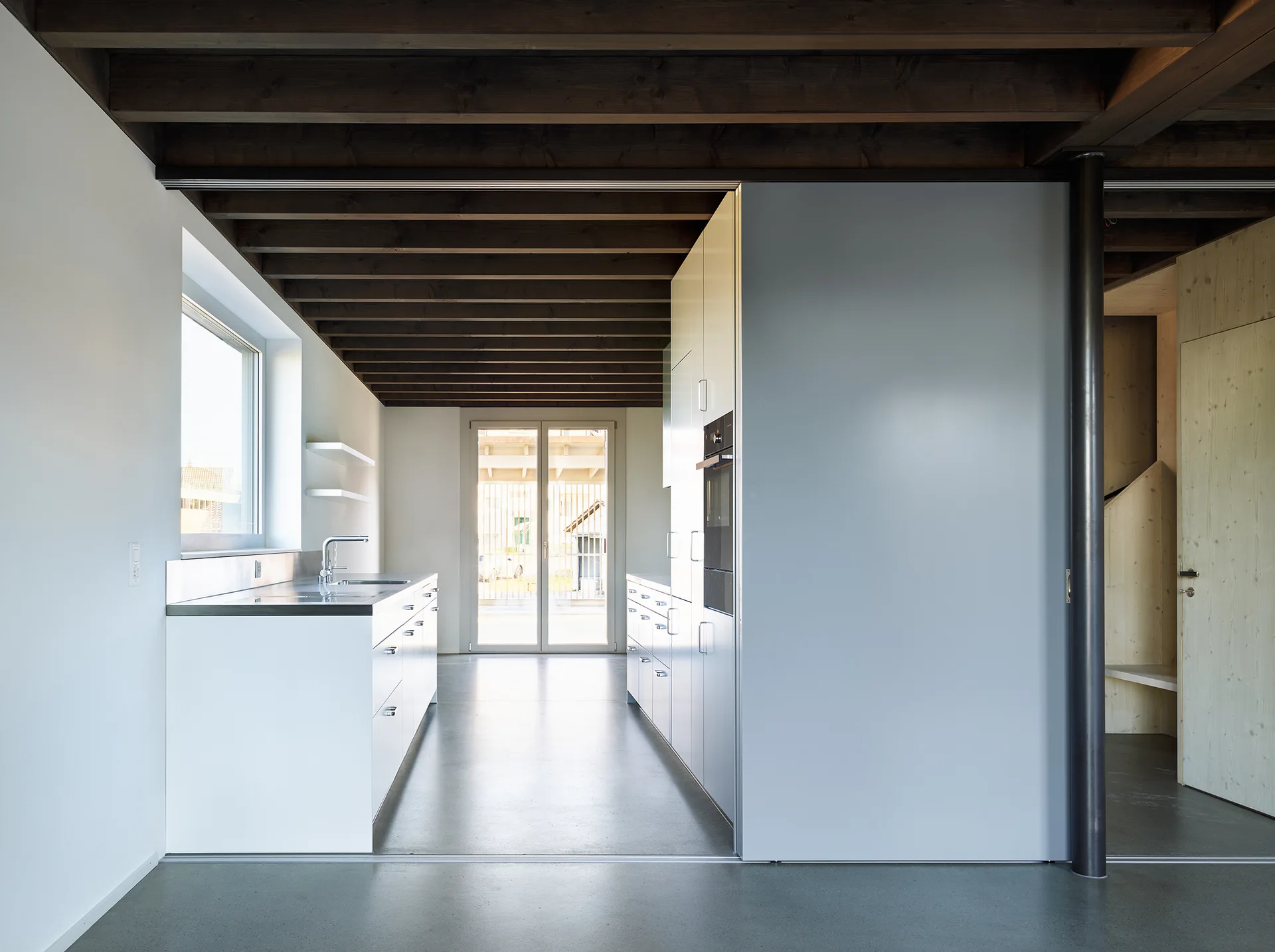 A modern galley kitchen features matte white cabinetry, stainless steel countertops, a grey concrete floor, and an exposed dark wood beamed ceiling.