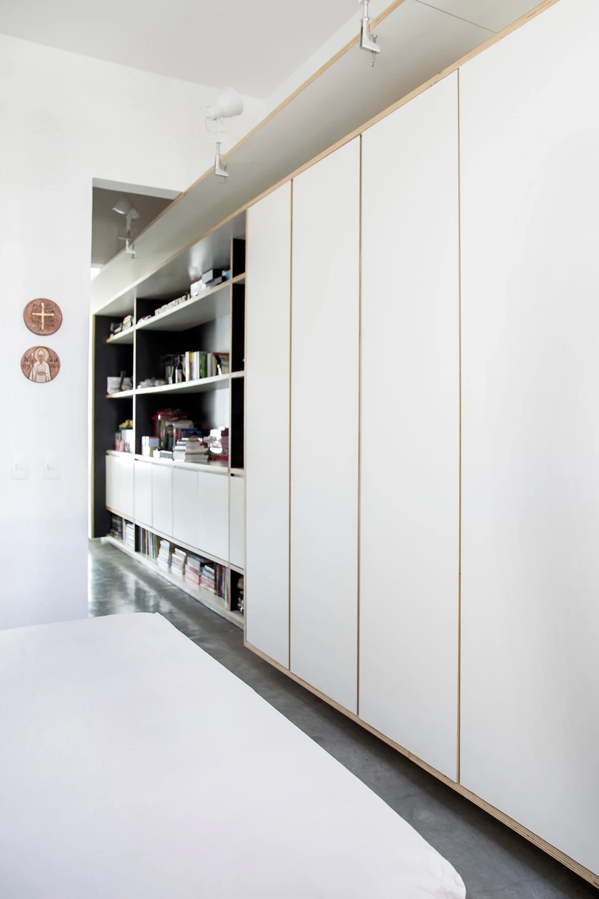 A bedroom features a large floor-to-ceiling white wardrobe with exposed plywood edges next to open built-in shelves filled with books.