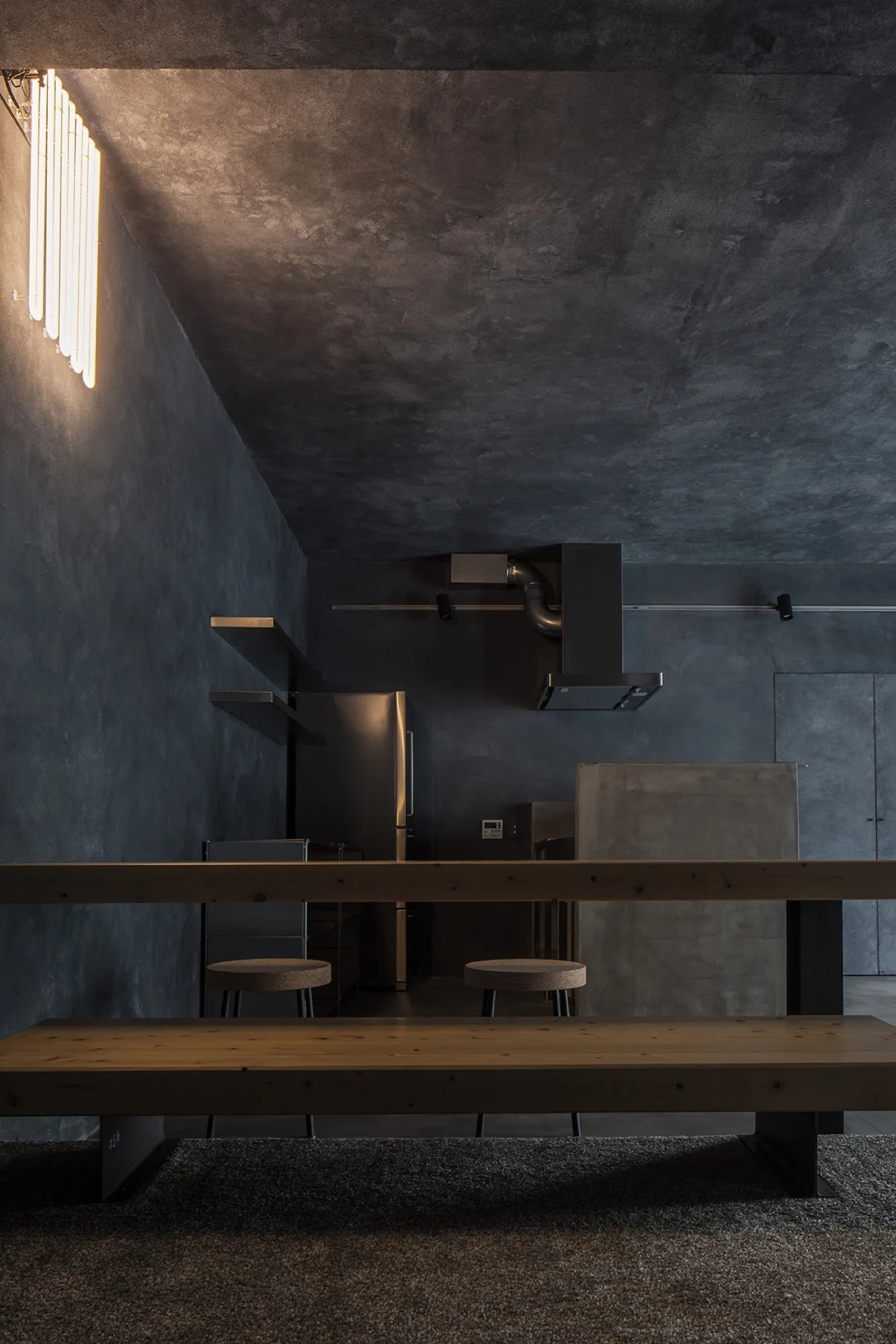 A dark industrial kitchen with textured charcoal walls, a concrete island, and a long wooden dining bench in the foreground.