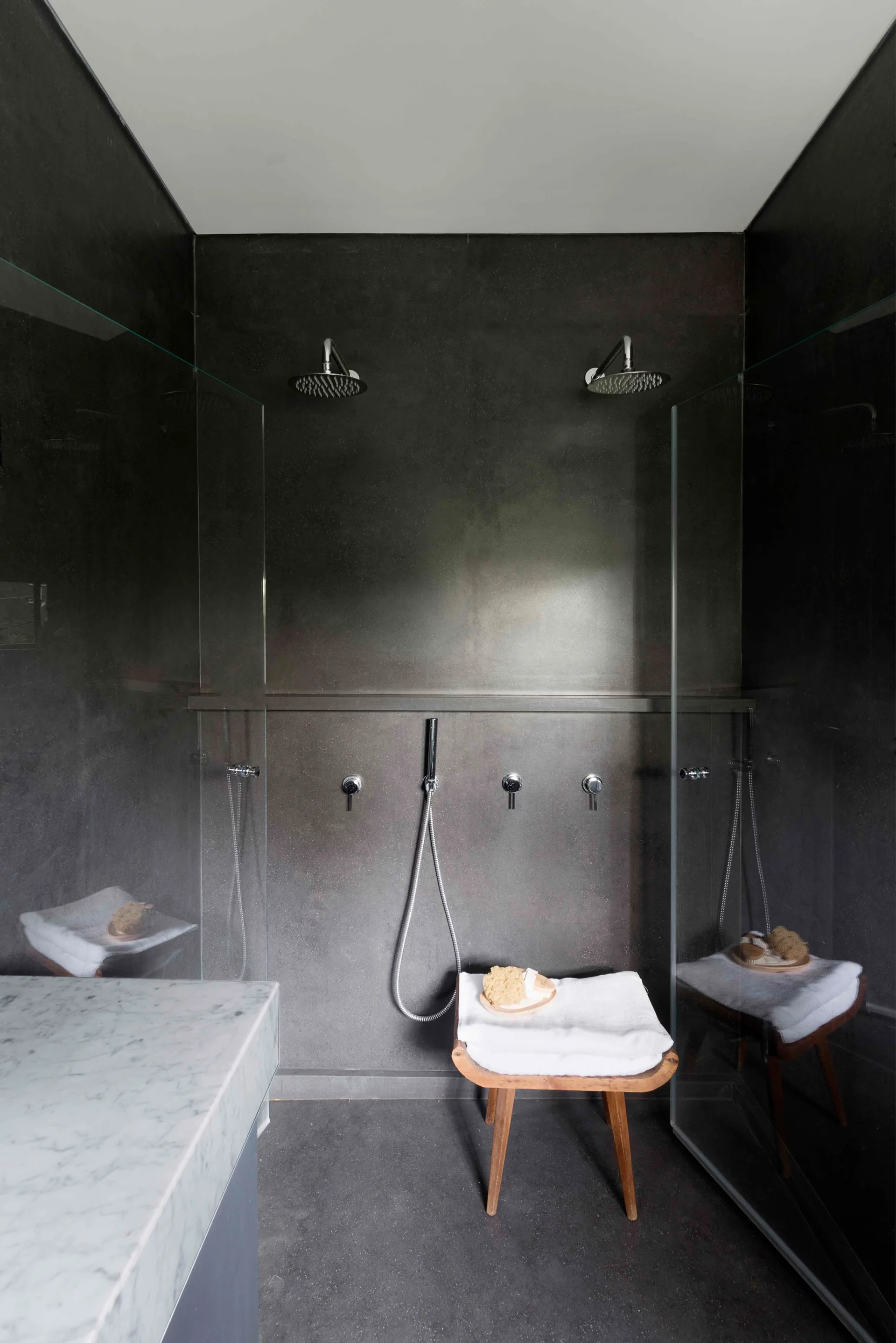 A minimalist walk-in shower features dark matte gray stone walls, dual chrome shower heads, glass partition panels, and a small wooden stool.