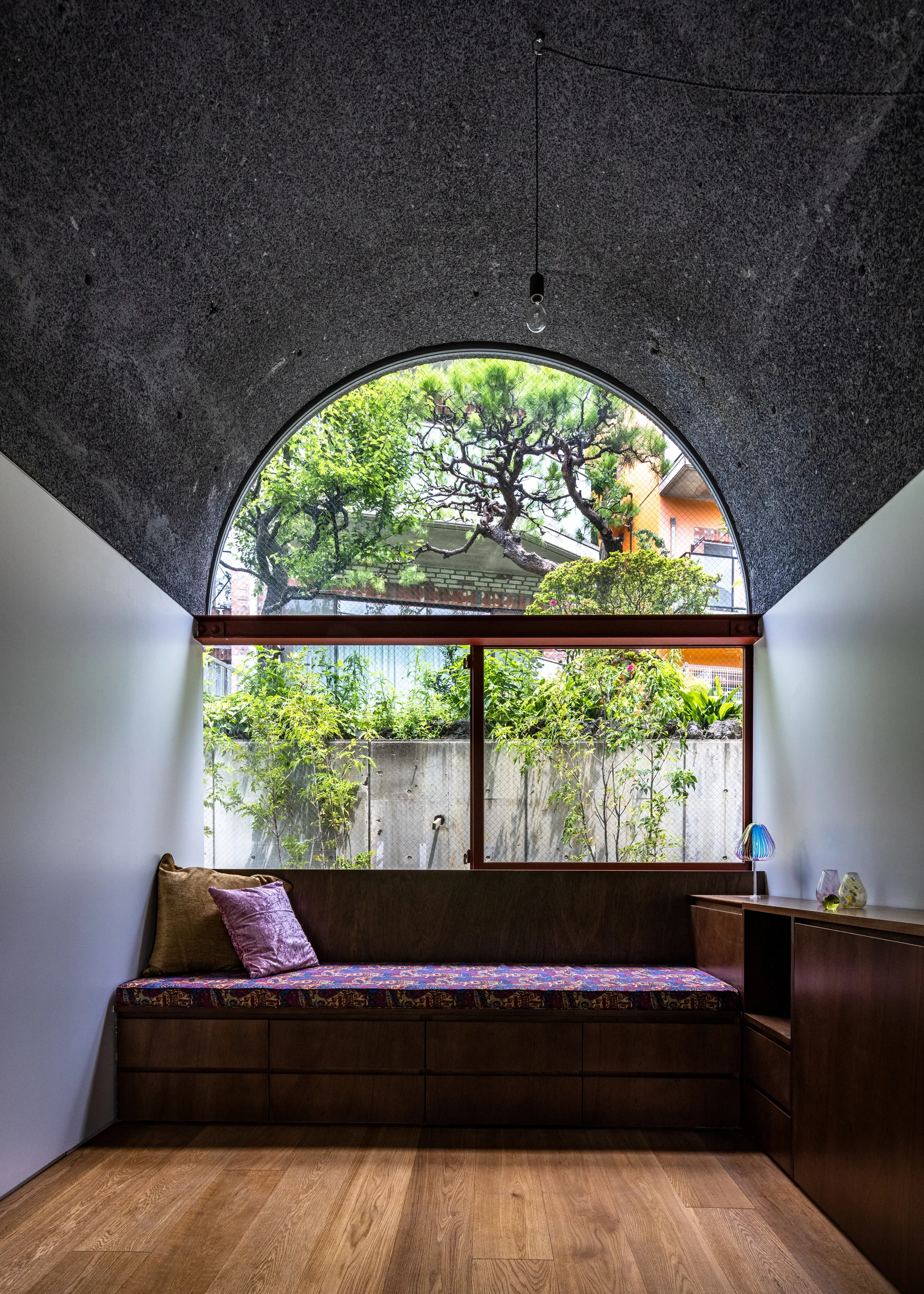A custom wooden storage bench with a patterned cushion sits beneath a large arched window looking out at a green garden.