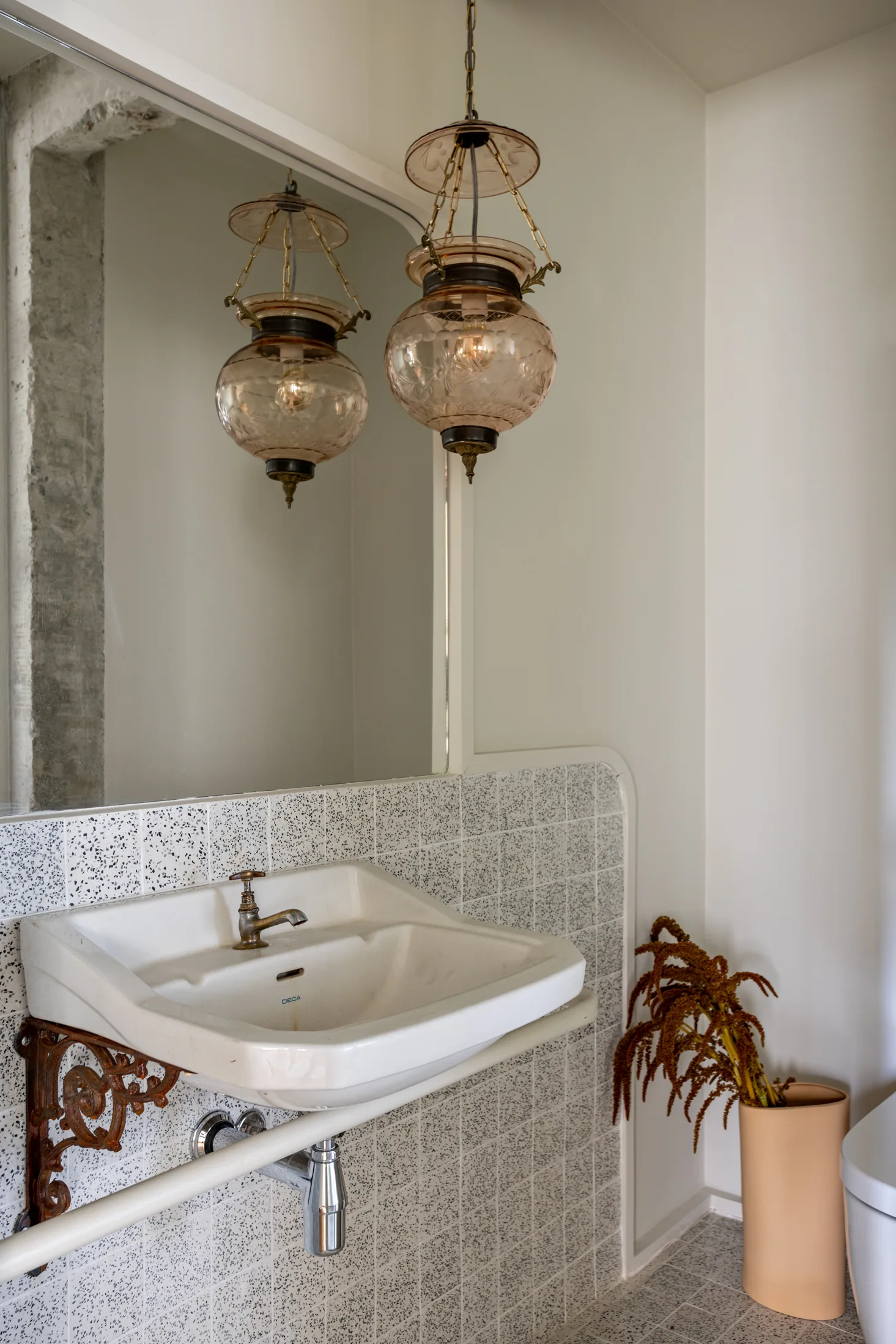 A wall-mounted white ceramic sink is supported by a rusted iron bracket in front of a speckled tile wall and an amber glass pendant lamp.
