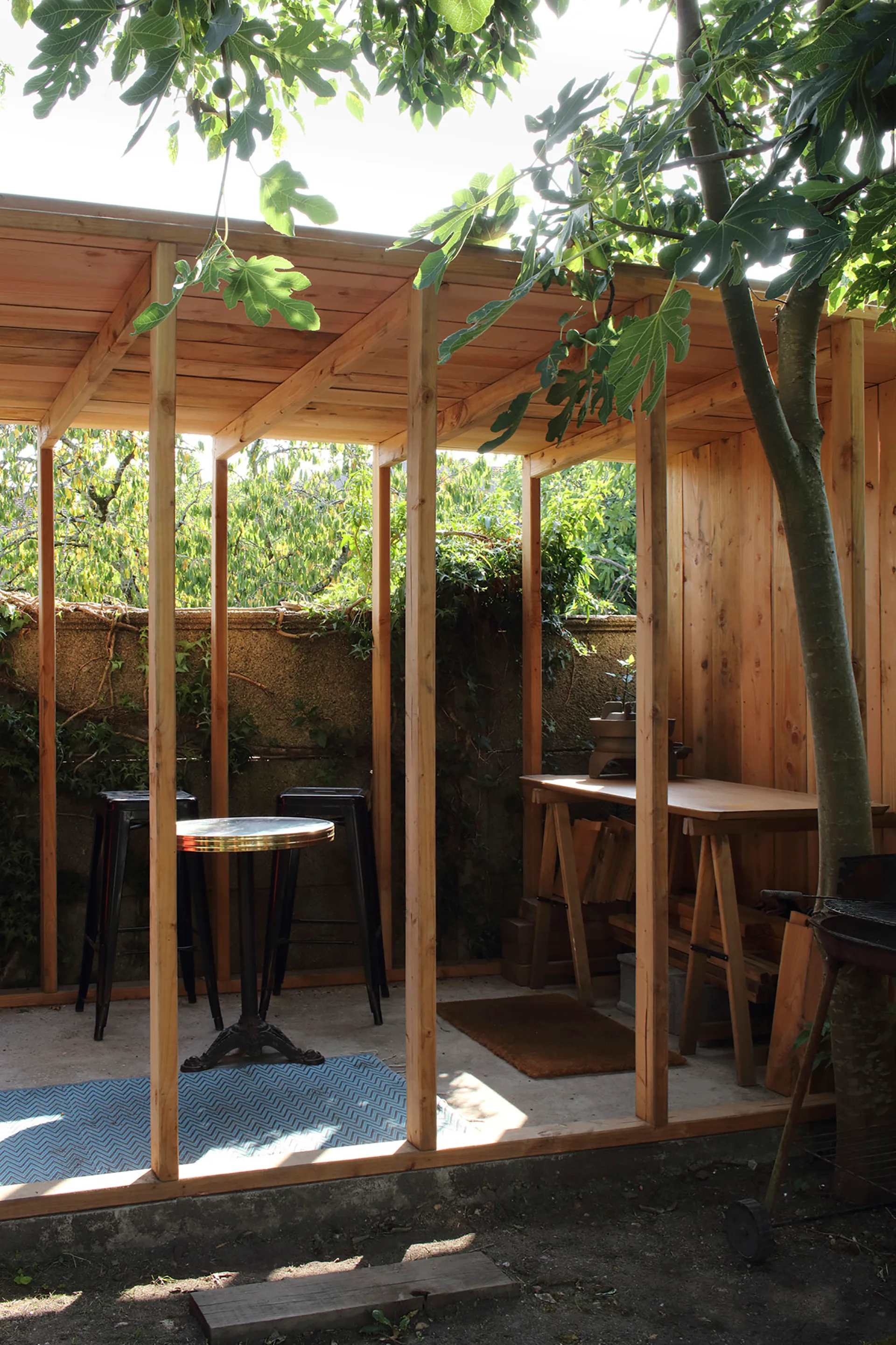A covered timber pergola on a concrete patio features a metal bistro table, black stools, and a wooden workbench under a green tree canopy.