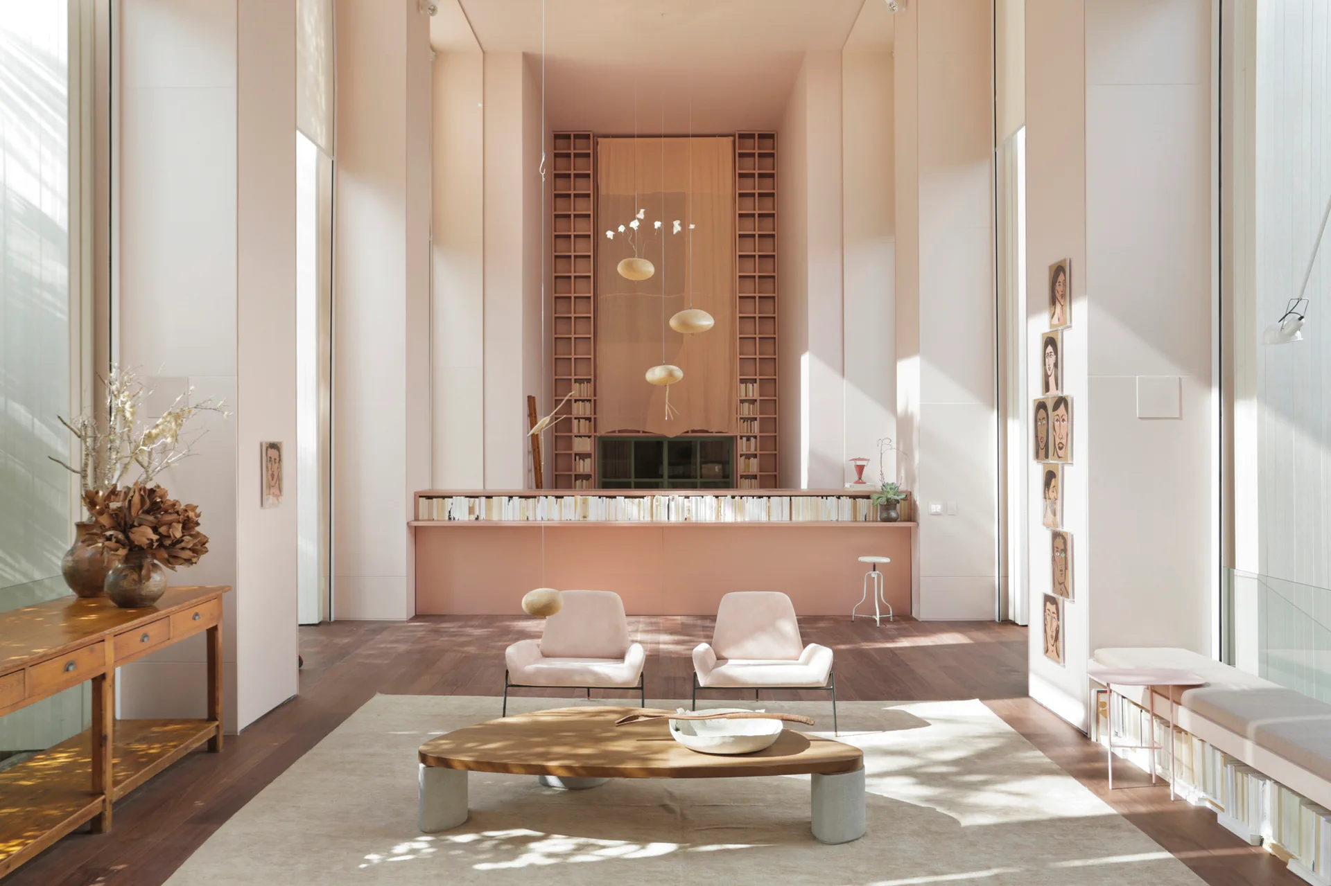 A spacious double-height living room featuring soft pink walls, two matching pink lounge chairs, a low wooden coffee table, and tall grid shelving.