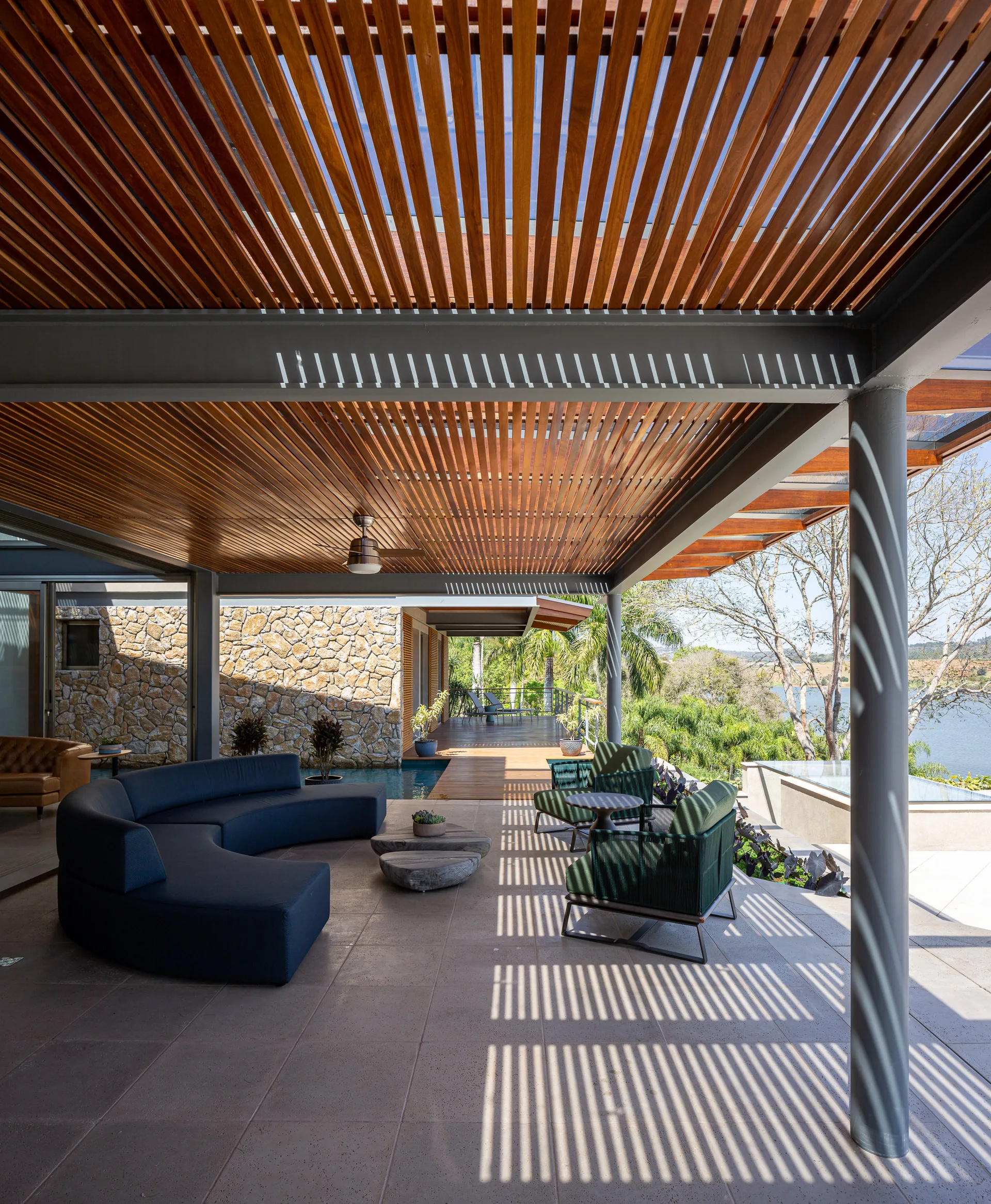 A covered outdoor patio features a curved dark blue sofa, green rope armchairs, and a slatted wood ceiling casting linear shadows.