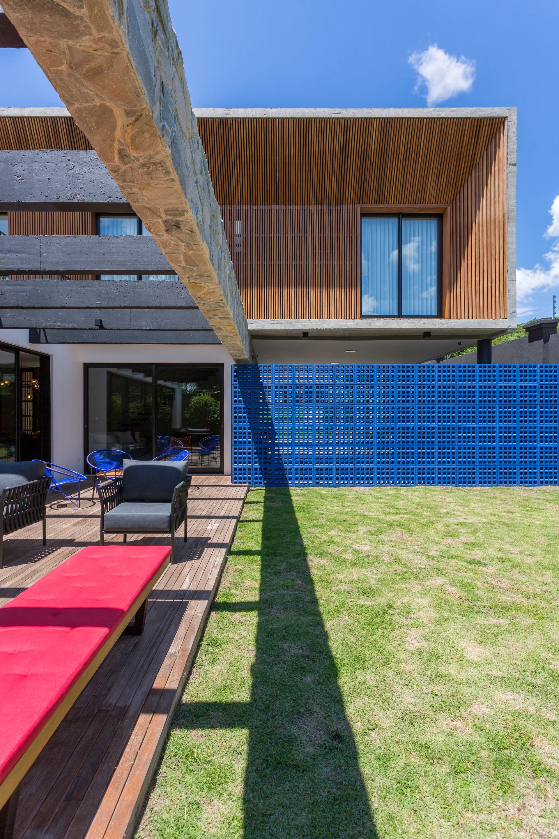 A wooden outdoor deck features dark grey lounge chairs, a bright red daybed, and a vivid blue breeze block privacy wall under a sunny sky.