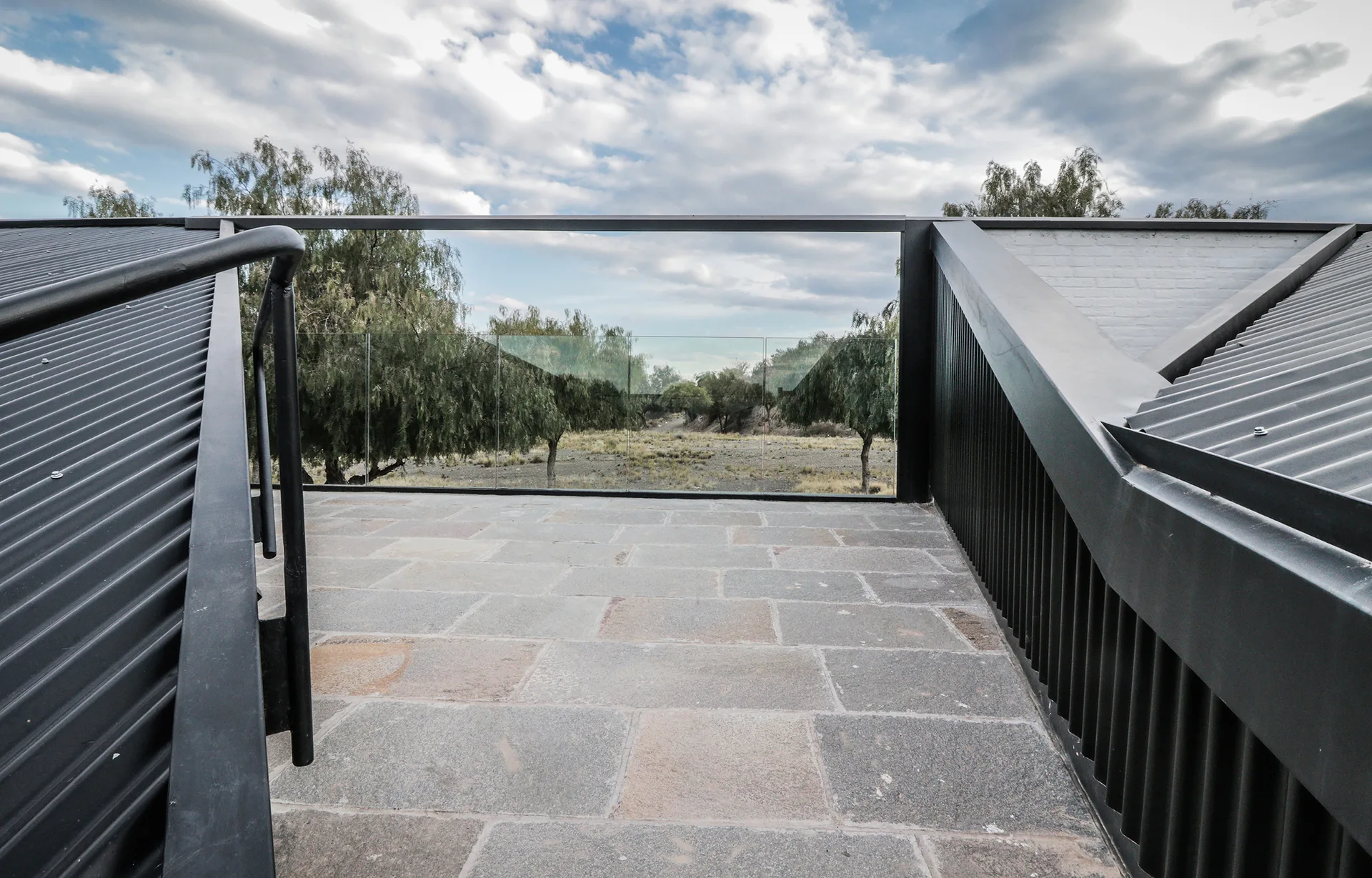An outdoor rooftop terrace features grey stone tile flooring, black corrugated metal roofing, and a clear glass railing overlooking trees.
