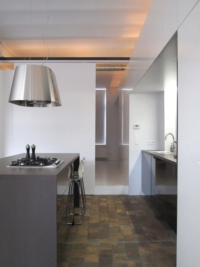 A minimalist kitchen features a dark grey island with a gas cooktop, a metallic range hood, and a rustic terracotta-tiled floor.