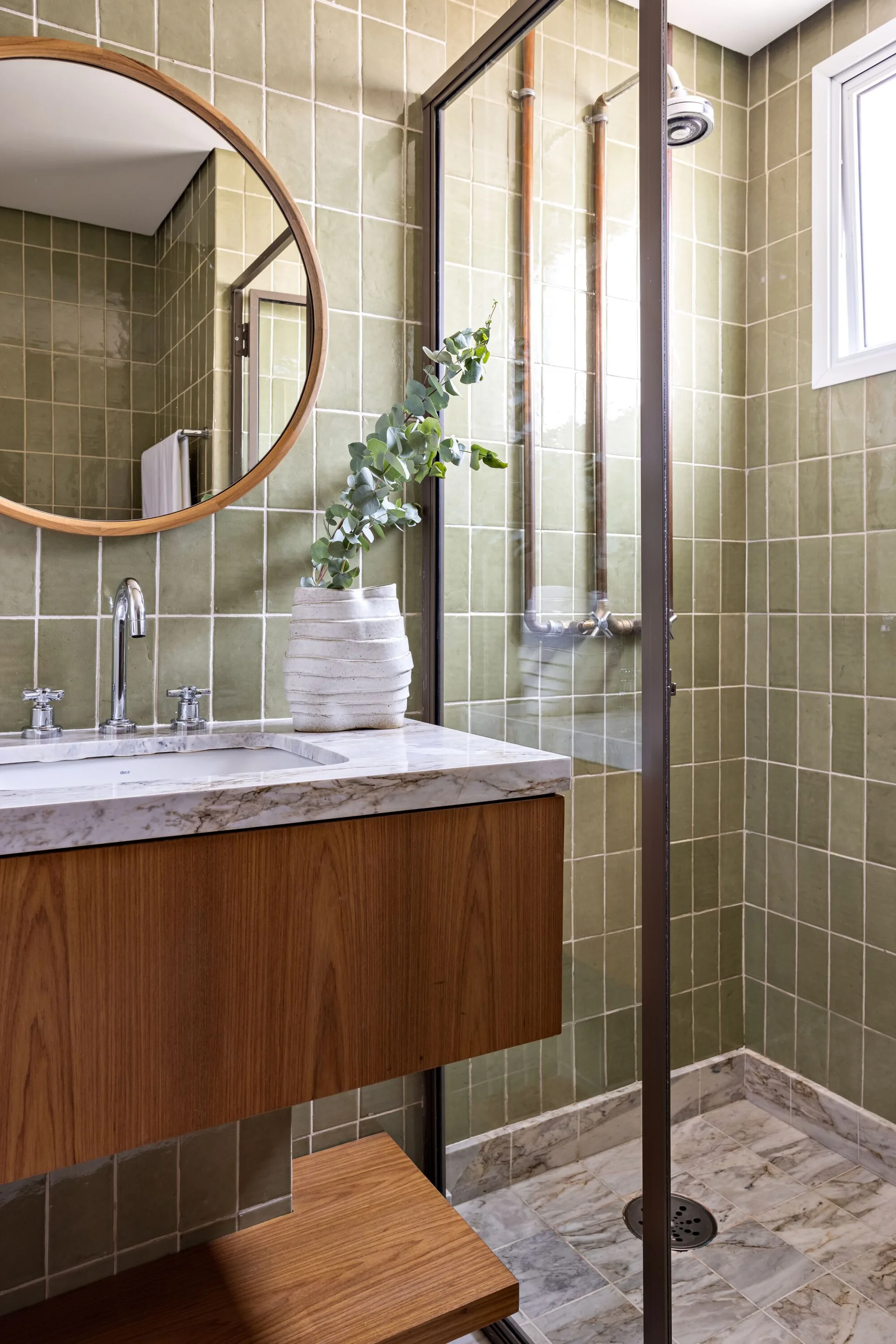 A bathroom with sage green square wall tiles, a wood vanity with marble top, a round mirror, and a glass-enclosed shower.