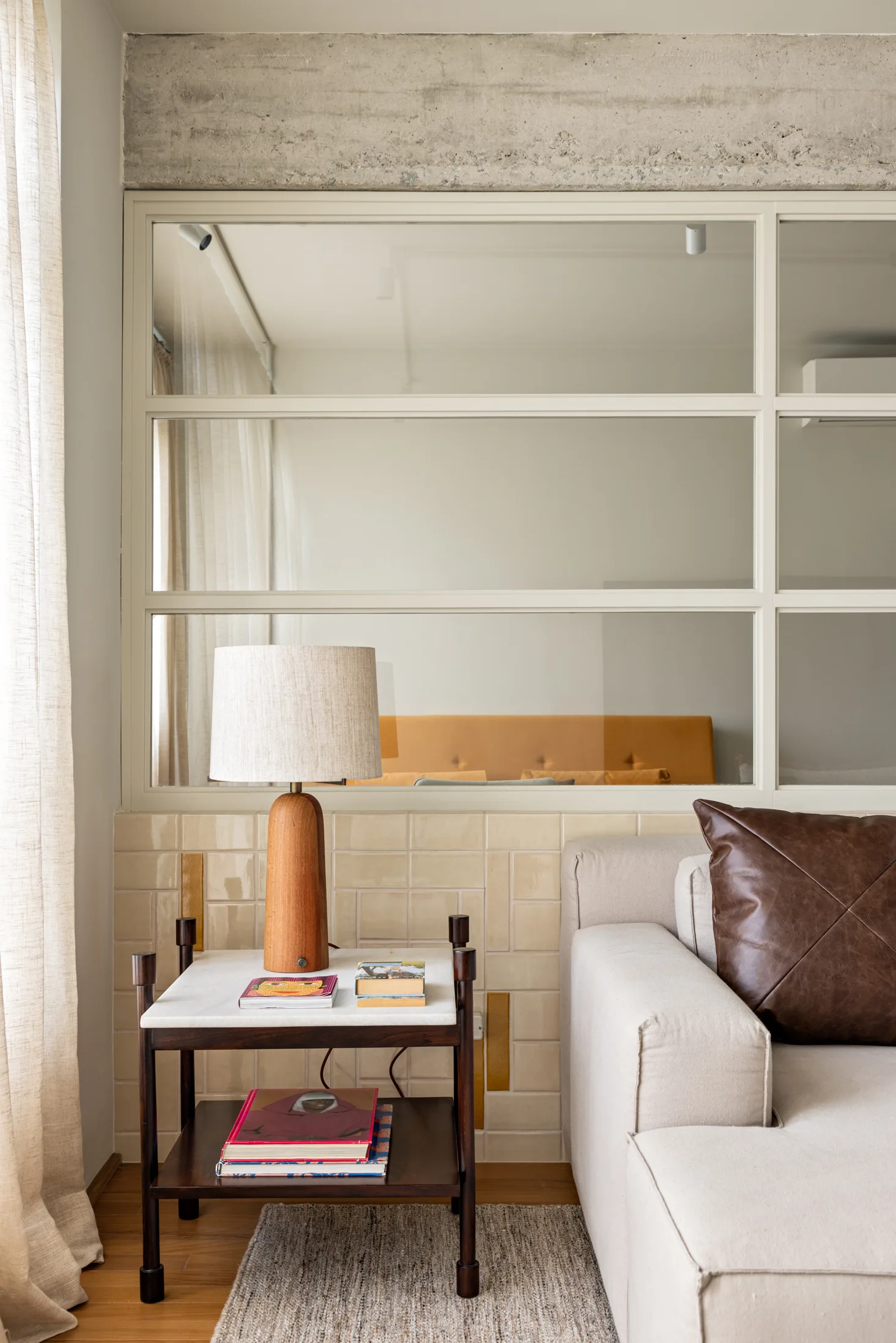 A beige sofa rests next to a two-tier wooden side table with a lamp, set against a cream tiled half-wall and an interior glass partition.